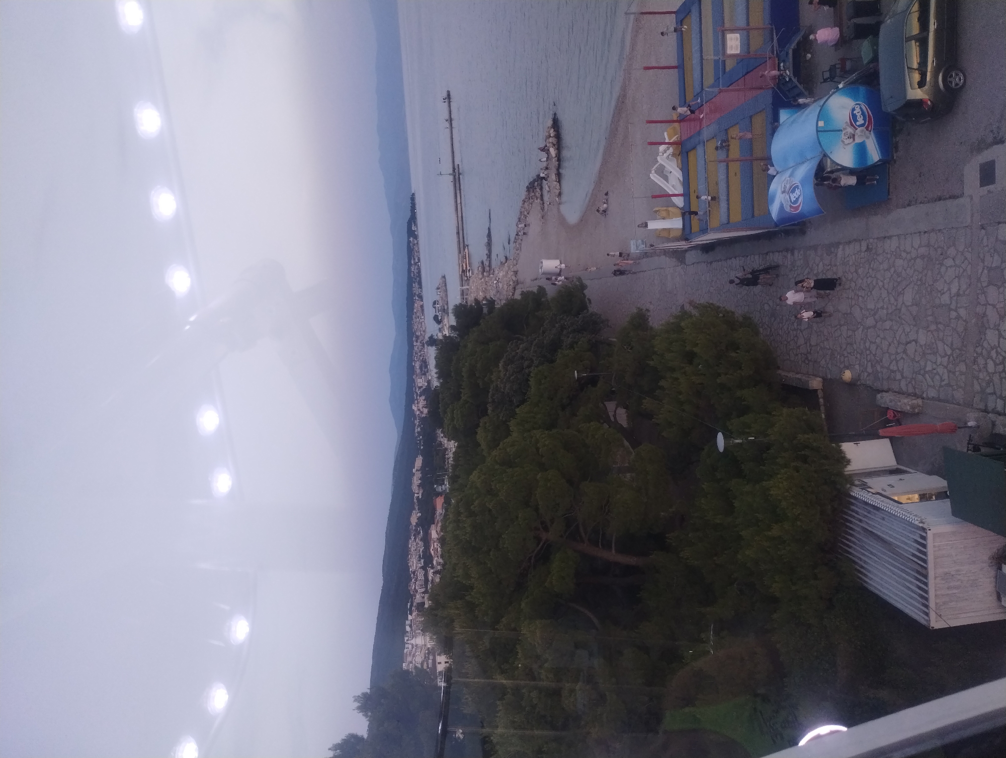 The image captures an aerial view of a coastal scene in Crikvenica, Croatia, likely during the late afternoon or early evening. The foreground is dominated by a paved walkway running alongside a beach. People are seen strolling along this walkway and on the beach, with some gathered on rocky outcrops along the shoreline.
Further down the beach, a children's entertainment area is visible, featuring trampolines and inflatable structures. Adjacent to this is a blue awning with branding that appears to be "Ledo," a Croatian ice cream and frozen food company. A dark-colored car is parked nearby.
The middle ground features a dense line of pine trees, separating the promenade and beach from a town that extends inland. The town consists of white and red-roofed buildings nestled against a backdrop of rolling hills.
In the distance, the Adriatic Sea stretches out to the horizon, with a pier extending into the water. The sky is overcast, creating a soft, diffused light. The top portion of the image shows a reflection, likely from glass, with several circular lights visible.