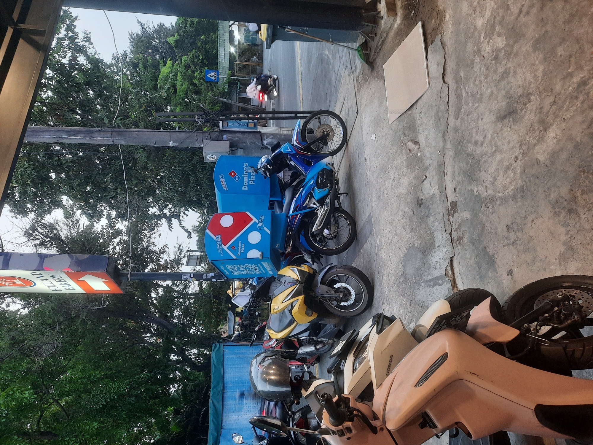The image depicts a street scene in North Jakarta, Indonesia, with a focus on parked motorcycles and a Domino's Pizza delivery box. Several motorcycles are parked on a concrete surface, some with helmets placed on them. In the background, a prominent sign for "HOLLAND BAKERY" is visible, pointing to the right. Adjacent to the bakery sign is a blue Domino's Pizza delivery box mounted on a motorcycle. Another Domino's Pizza delivery motorcycle is parked further down the street. The environment appears to be urban, with trees lining the street and a pedestrian crossing visible in the distance, where a person on a motorcycle is passing by. The overall impression is of a typical commercial area in a city, with active delivery services.