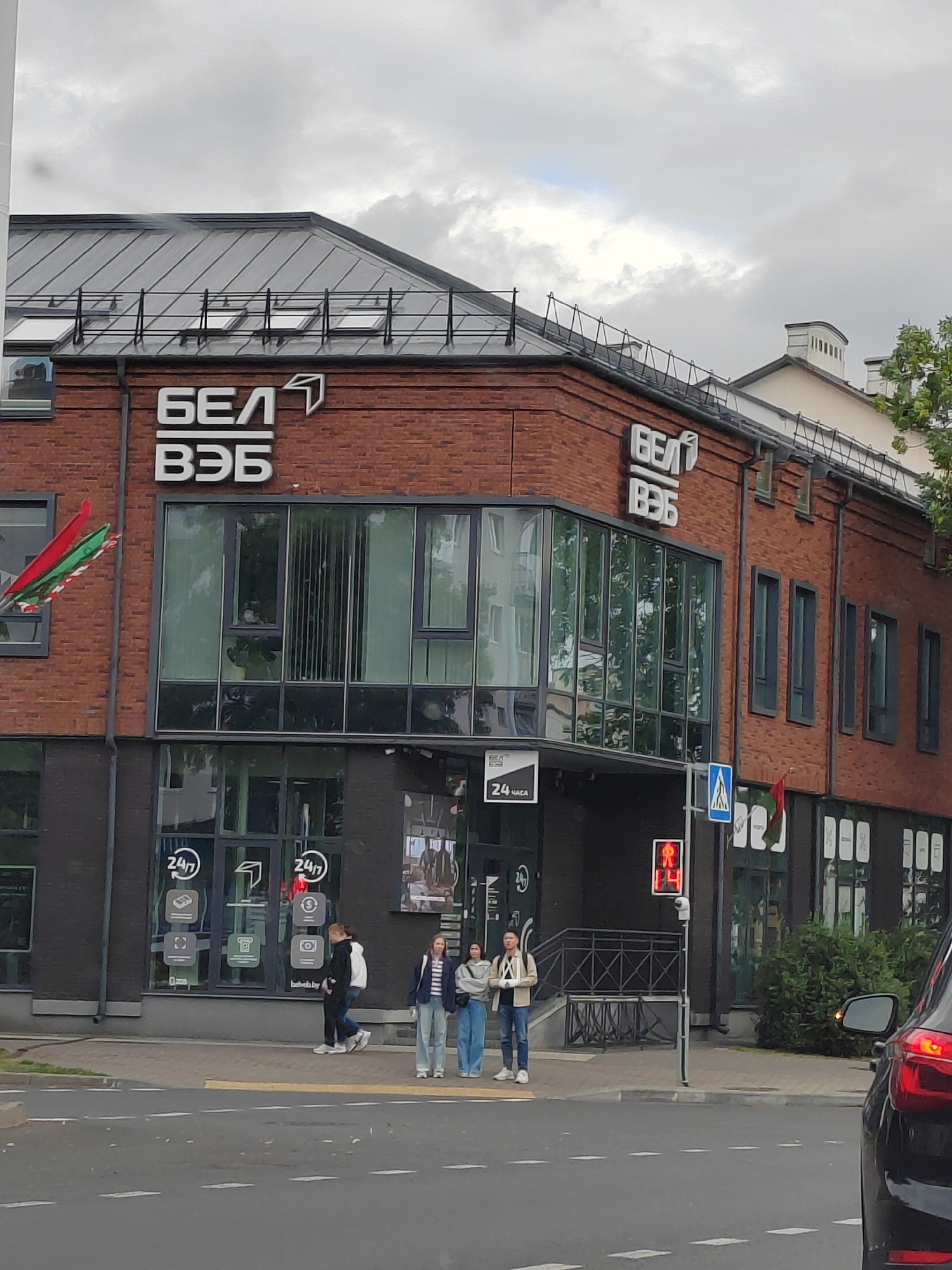 The image shows a modern brick building with large glass windows and a metal roof. The building has prominent white signage on its facade that reads "БЕЛ" and "ВЭБ". A red, green, and white flag is visible on the left side of the building. In front of the building, there is a sidewalk with several people walking and standing. A traffic light displaying a red pedestrian signal is visible on the right side of the image, along with a street sign. The street in front of the building has white lane markings. The sky is overcast. This building is likely located in the city of Brest, Belarus, as indicated by the presence of Belarusian language on the signage and the national flag.