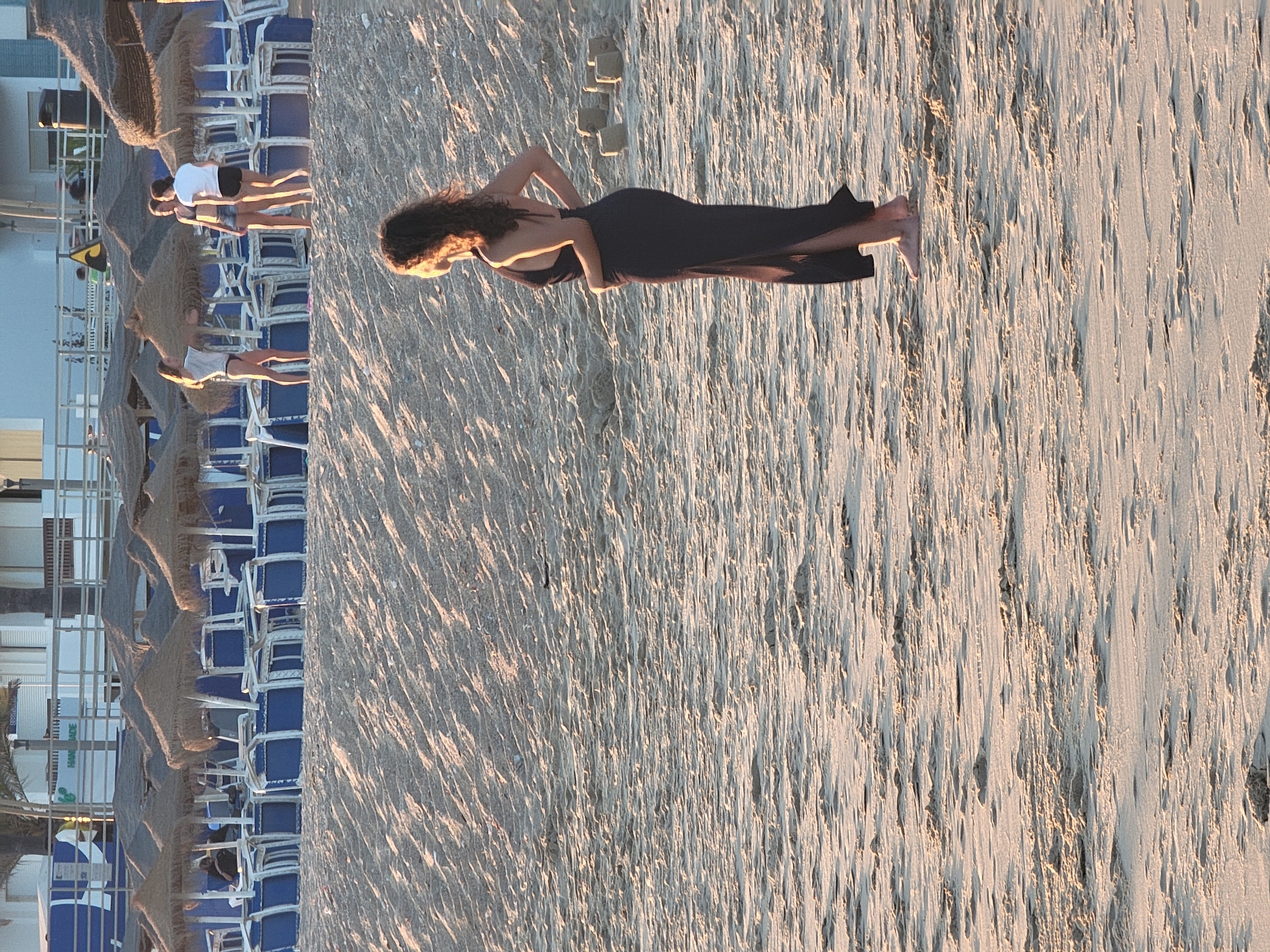 The image depicts a sandy beach in Quarteira, Portugal, likely during the late afternoon or early evening given the warm, low-angled lighting casting long shadows across the sand. In the foreground, a woman with long, curly dark hair is standing, facing away from the camera and looking out towards the ocean. She is wearing a long, dark, sleeveless dress with a halter neck. Her hands are resting on her hips, and her body is angled slightly to her left. Her bare feet are visible on the sand.
In the mid-ground, behind a ridge of sand, a row of beach umbrellas with thatched roofs stands over stacked blue lounge chairs. Several people are visible in this area, though their details are less clear due to distance and lighting. Three young women are standing in a group, appearing to be in conversation. One woman in the front is wearing a white t-shirt and dark bottoms, another in the middle is wearing a dark bikini top and denim shorts, and the third, further back, is wearing a white t-shirt and dark shorts.
The background shows a building structure with balconies and railings, suggesting a hotel or resort adjacent to the beach. The sand itself is textured with ripples and small mounds, indicative of wind and wave action. Scattered on the sand are several small, cuboid structures that appear to be sandcastles or sand sculptures. The overall impression is one of a peaceful beach scene at the end of the day.