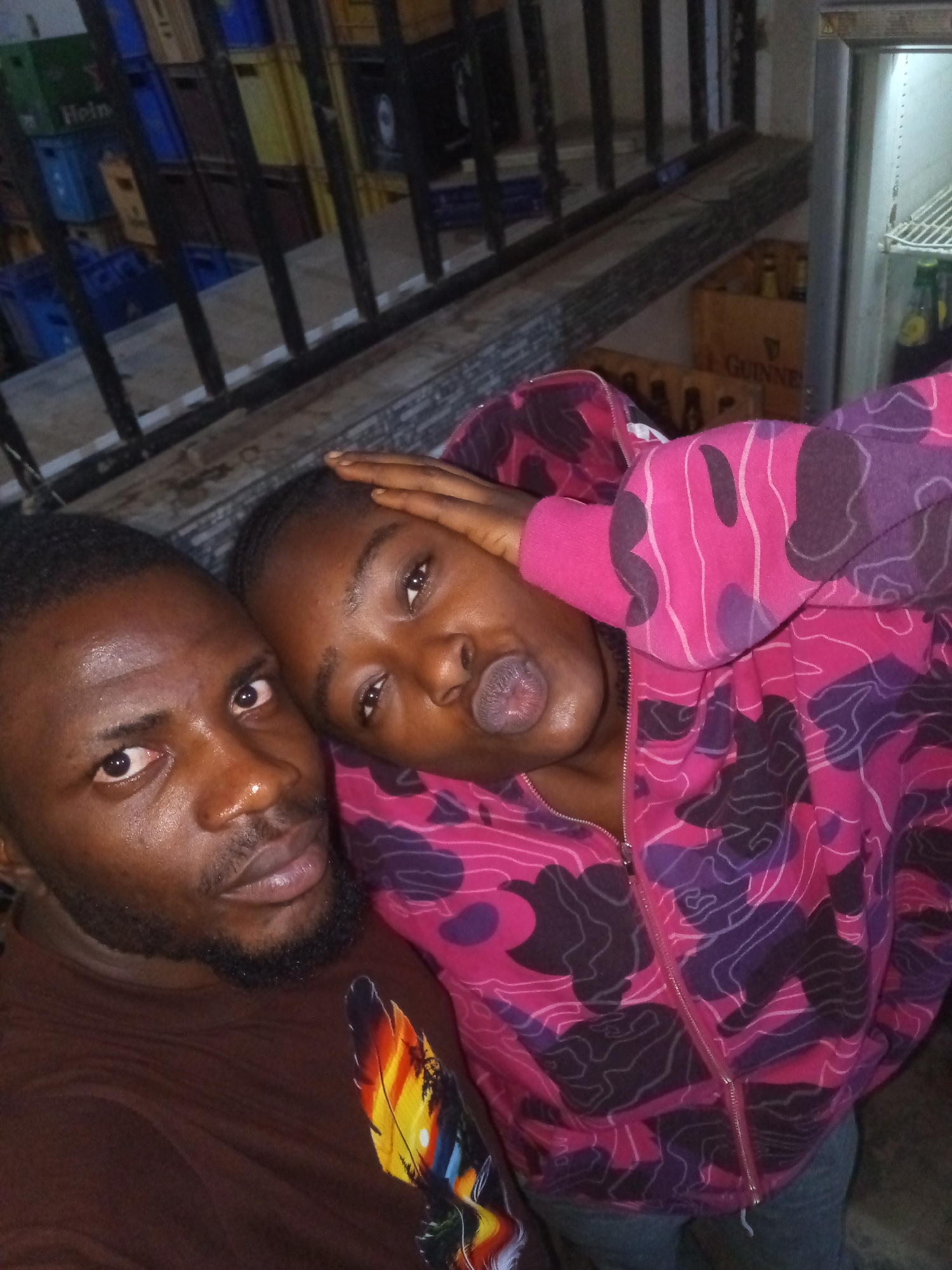 Two people, a man and a woman, are in the foreground of the image, posing for a selfie. The man is on the left, facing the camera with his left hand on his beard. He is wearing a brown t-shirt with a colorful feather design on the left side. The woman is on the right, leaning her head on the man's shoulder and holding her right hand on her forehead. She is wearing a pink hooded jacket with a camouflage pattern in black and purple. Her lips are pursed. Behind them, there are stacked crates and a refrigerator. The environment appears to be a storage area or a small shop. The lighting suggests an indoor setting.