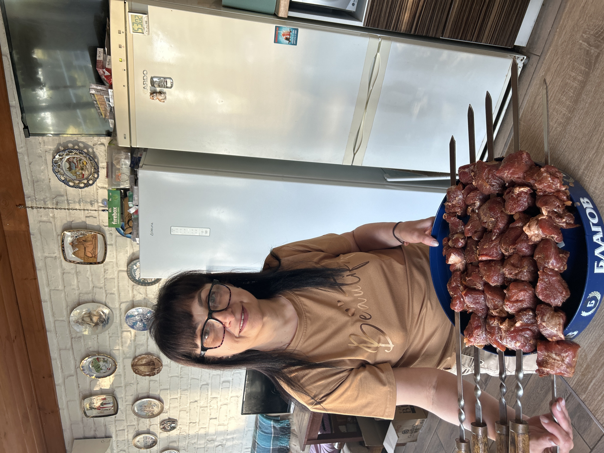 A woman with dark hair and glasses stands in a room with white brick walls, holding a blue tray filled with raw meat skewers. She is wearing a tan t-shirt with the word "Beautiful" printed on it. Behind her, two refrigerators are visible, one white and one lighter white. A television is mounted on the wall above the refrigerators. Decorative plates are displayed on the brick wall. The room appears to be a kitchen or dining area, possibly in Hnarovske, Ukraine, given the context.