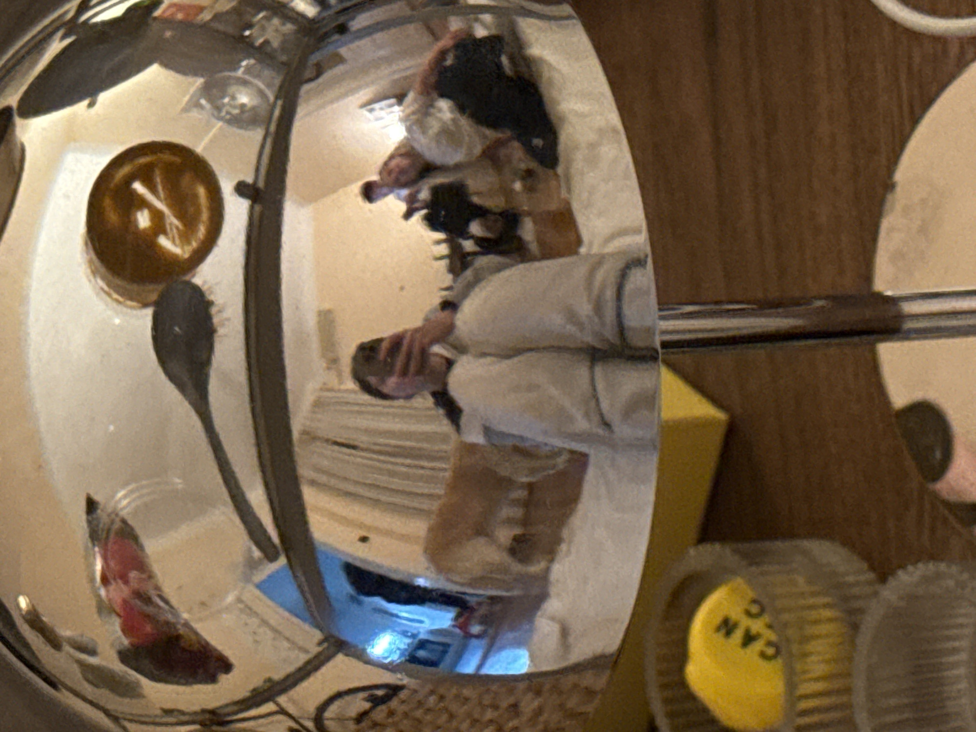 The image displays a highly distorted reflection captured within a shiny, curved metallic object, likely a pot or kettle, resting on a wooden surface. The reflection provides a wide, fisheye-like view of an indoor scene, presumably a room in a residential setting.
In the reflected view, a person is prominently visible, wearing light-colored attire, possibly a hoodie or sweatshirt. They appear to be looking downwards, their hands occupied with an object, likely a mobile phone or tablet, which partially obscures their face. Beside and behind this person, a smaller figure, possibly a pet or a child, can be seen lying or sitting on a surface that resembles a bed or sofa.
Other reflected objects include a dark, circular item, possibly a cup or bowl, containing a lighter stick-like object such as a spoon or stirrer. Adjacent to this is a dark hairbrush. Further down in the reflection, a device emitting a blue light is discernible on a lower surface, accompanied by what appears to be an electrical cable. The background of the reflection shows light-colored walls and a ceiling light fixture.
Outside the main reflective surface, a rich brown, grainy wooden texture, characteristic of a table or countertop, occupies the right side of the frame. At the bottom right, a yellow, circular cap is visible, featuring the partial text "CAN C". Nearby are clear, ribbed plastic components, suggesting parts of a kitchen appliance such as a food processor or blender. The overall lighting suggests a well-lit indoor environment, either during the day or with bright artificial lighting. Due to the significant distortion, emotions are not discernible, and no specific location cues for Belgrade are visible within the image itself.