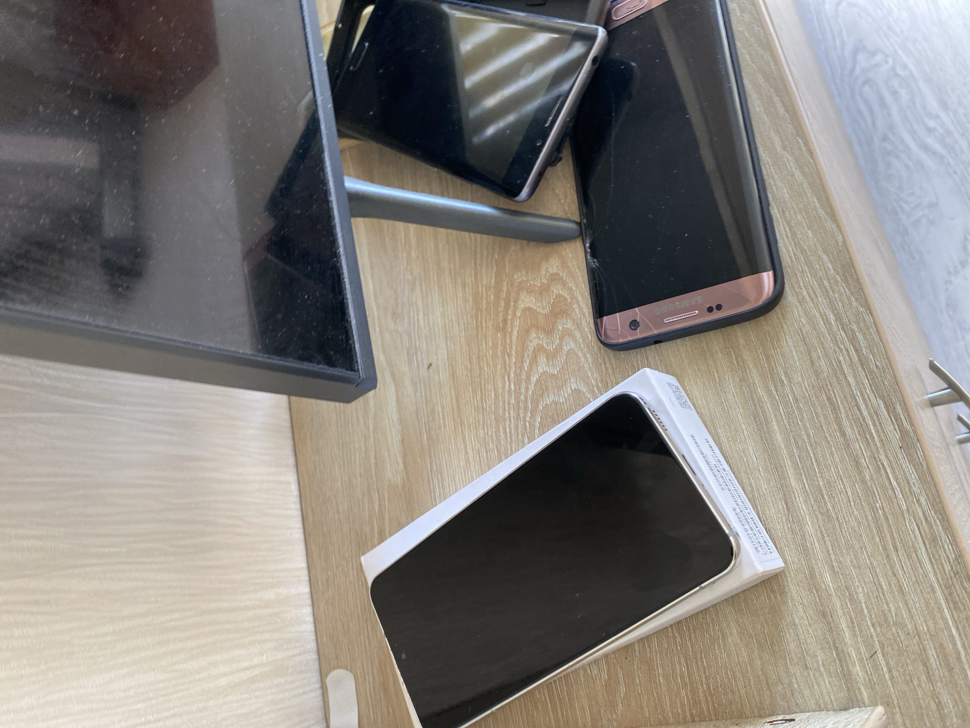 The image shows a shelf with several electronic devices. On the left, a white box contains a black smartphone with a silver frame. To the right of the box, another black smartphone with a rose gold frame and a cracked screen is visible. Behind these, a tablet with a black screen is partially obscured. Above these devices, a dusty flat-screen monitor is positioned. The background consists of a light-colored, textured wall. The scene appears to be an indoor setting, possibly a home or office, on a wooden surface. No specific location in Chișinău, Moldova, can be determined from the image.