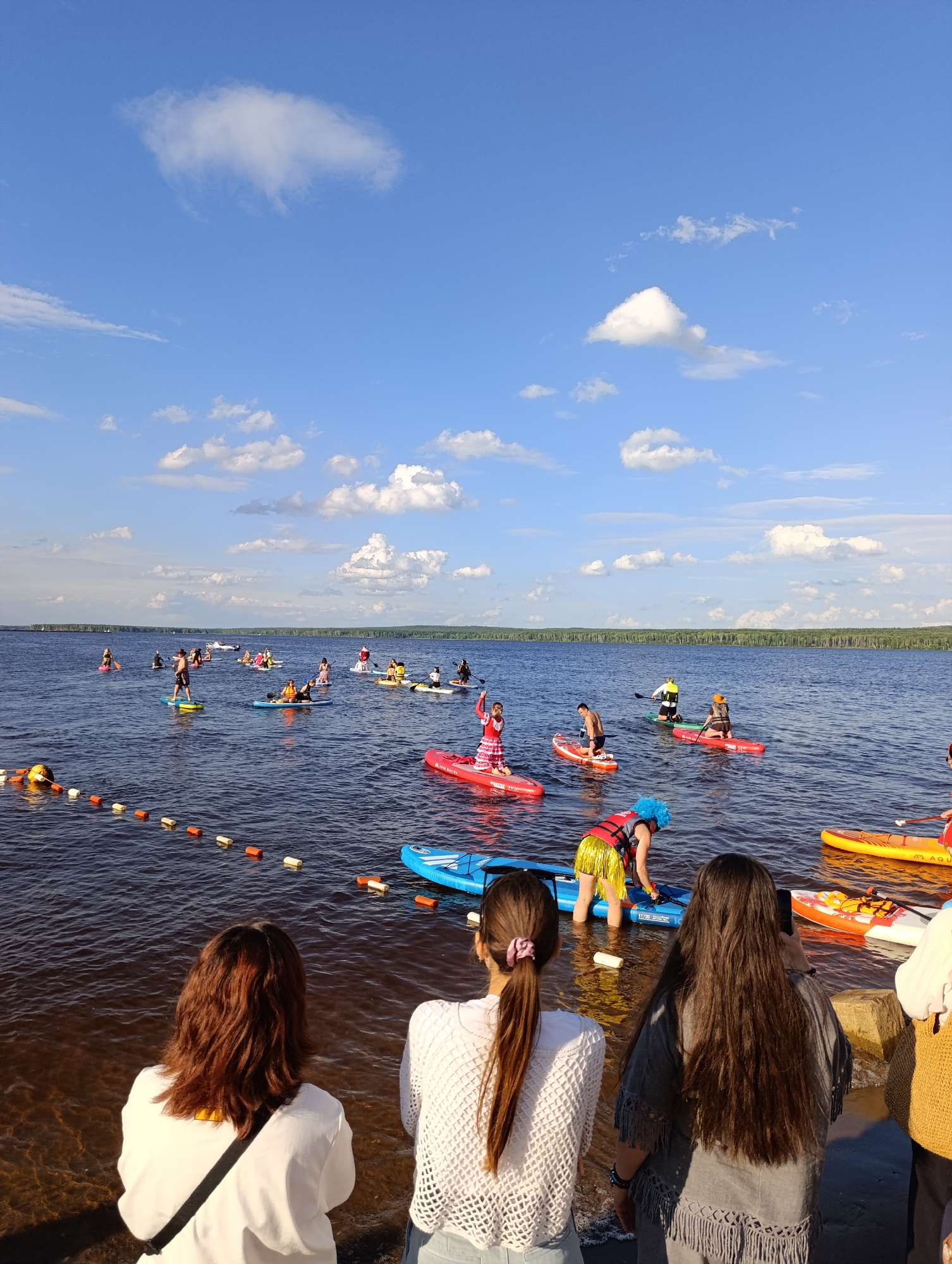 This is a wide shot of a lake with many people paddleboarding. The subjects are a diverse group of individuals on paddleboards, scattered across the water. Objects include the paddleboards themselves, some with paddles. The scene is a large, calm body of water under a bright blue sky with fluffy white clouds. The setting is a lake, likely in Chaikovsky, Russia, with a treelined shore visible in the distance.
The activity taking place is a paddleboarding event or gathering. People are paddling, standing, and some are kneeling on their boards. It appears to be a recreational event, possibly a group excursion or a competition.
Notable details include the time of day, which appears to be late afternoon or early evening, given the quality of light and shadows. The weather is clear and pleasant, with a bright sun and a blue sky. The emotion conveyed is one of fun and enjoyment, with participants engaged in a shared outdoor activity.
There are a few visible text elements on some of the paddleboards, but they are too small and blurry to be legible. A rope with orange and white buoys is visible in the foreground, marking off an area of the water.