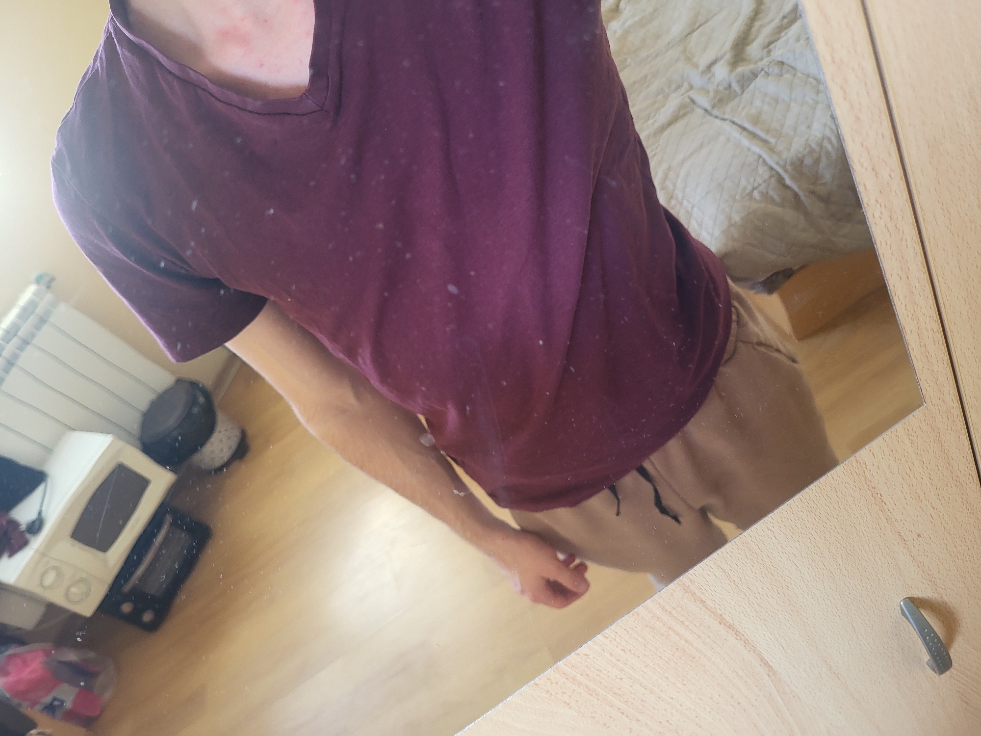 The image shows a reflection in a mirror of a person wearing a purple t-shirt and khaki pants. The t-shirt is visibly worn with lighter-colored spots. In the background, reflected in the mirror, are elements of a room including a radiator, a small microwave, and a bed with a light-colored comforter. The background objects suggest the room is likely in a domestic setting, potentially an apartment or house. The image does not contain information suggesting the location is Sofia, Bulgaria.