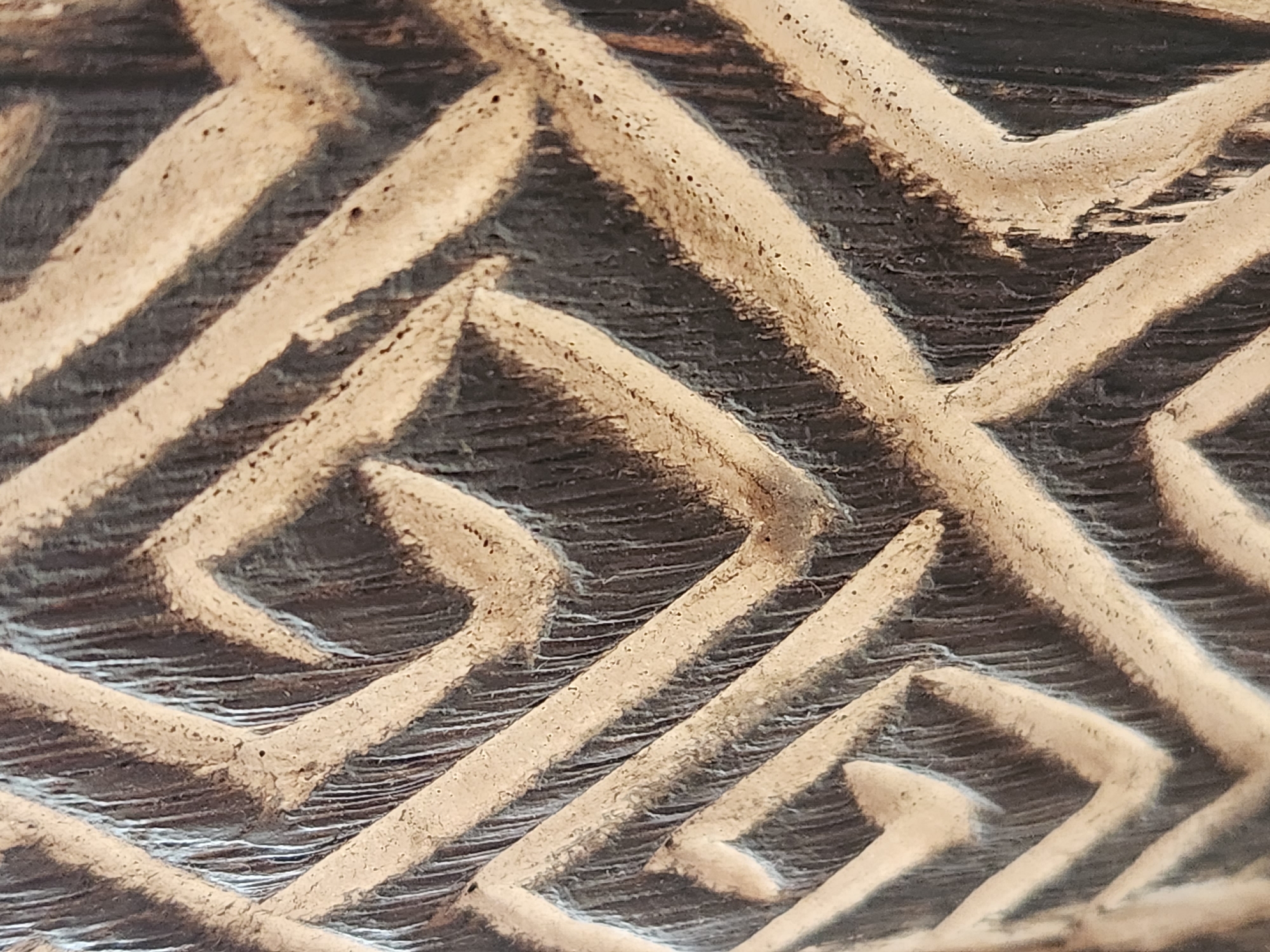 The image shows a close-up, detailed view of an abstract, carved pattern on a dark, textured surface, possibly wood. The pattern consists of intersecting diagonal lines and geometric shapes, creating a maze-like or labyrinthine effect. The carved areas are lighter in color and have a rough, granular texture, contrasting with the darker, smoother background that appears to have vertical striations. The lighting casts shadows that accentuate the three-dimensional depth of the carving. There are no people, animals, or identifiable objects present, and the context of its location in Maitland, Florida, United States of America, is not visually evident from the image itself.