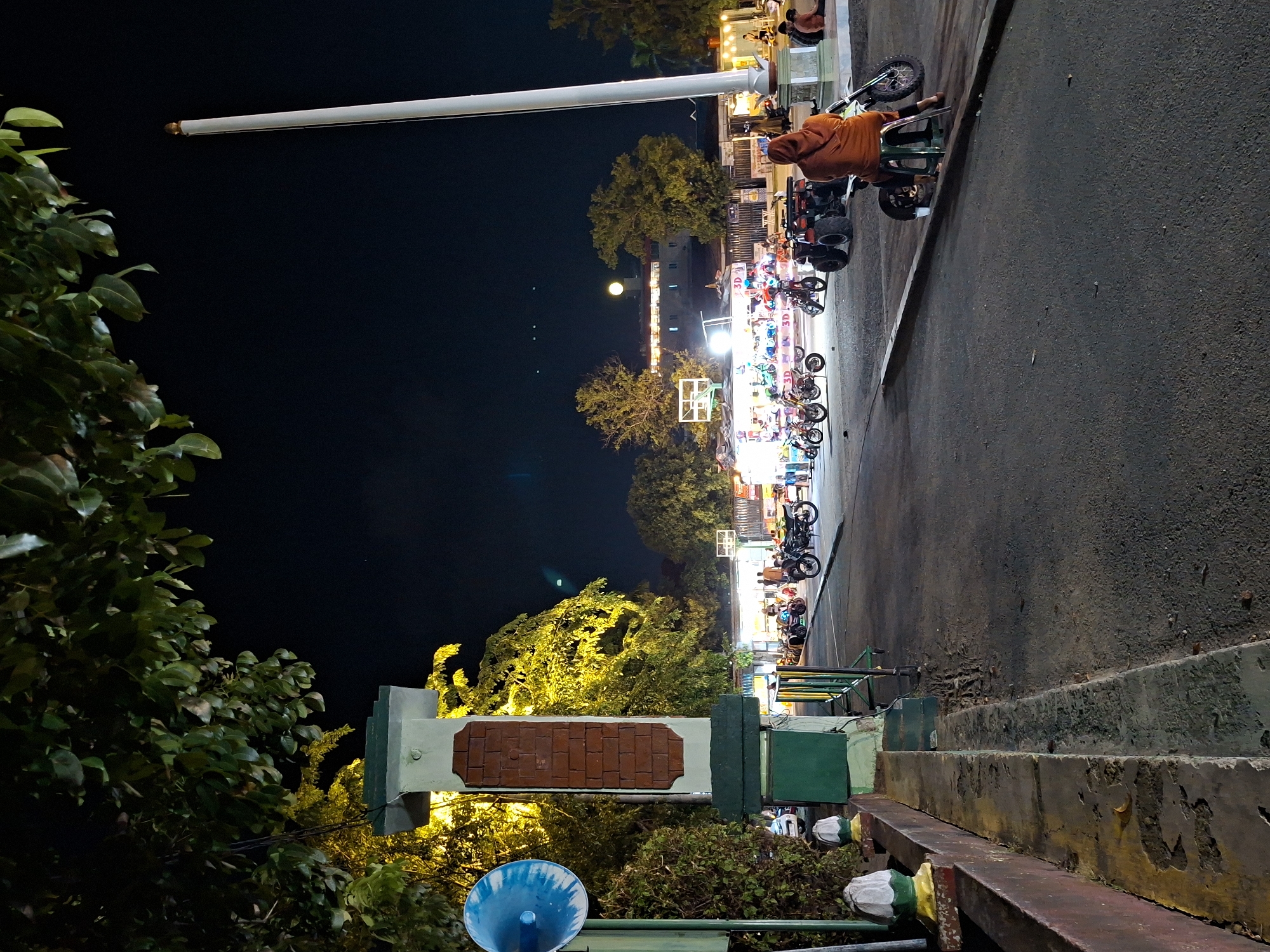 This nocturnal photograph captures an illuminated outdoor recreational area in Lhokseumawe, Indonesia. The scene is set under a dark night sky, where a crescent moon is subtly visible.
On the left side of the frame, a tall, slender white flagpole extends upwards, partially obscured by lush green foliage in the foreground. Further back, several trees with yellowish-green leaves are brightly lit by artificial sources, suggesting decorative lighting.
The right side of the image features a dark grey paved road or pathway, bordered by concrete steps and low walls. Along this pathway, a line of small recreational vehicles, including mini motorcycles, quad bikes, and scooters, is neatly arranged. These vehicles are brightly lit by colorful lights, and some display "3D" text, indicating a possible rental business or attraction. A person wearing an orange hooded top is seated on one of these vehicles, possibly waiting or resting. In the background, more people are visible near distant, illuminated buildings, further enhancing the lively evening atmosphere.
In the lower foreground, more dense green foliage is present, alongside a distinctive structure featuring a brick-patterned section atop a green base. A circular blue object, possibly a speaker or part of a decorative fountain, is also visible near the bottom left. The overall impression is that of a bustling local night market or park area, offering leisure activities under artificial light.
