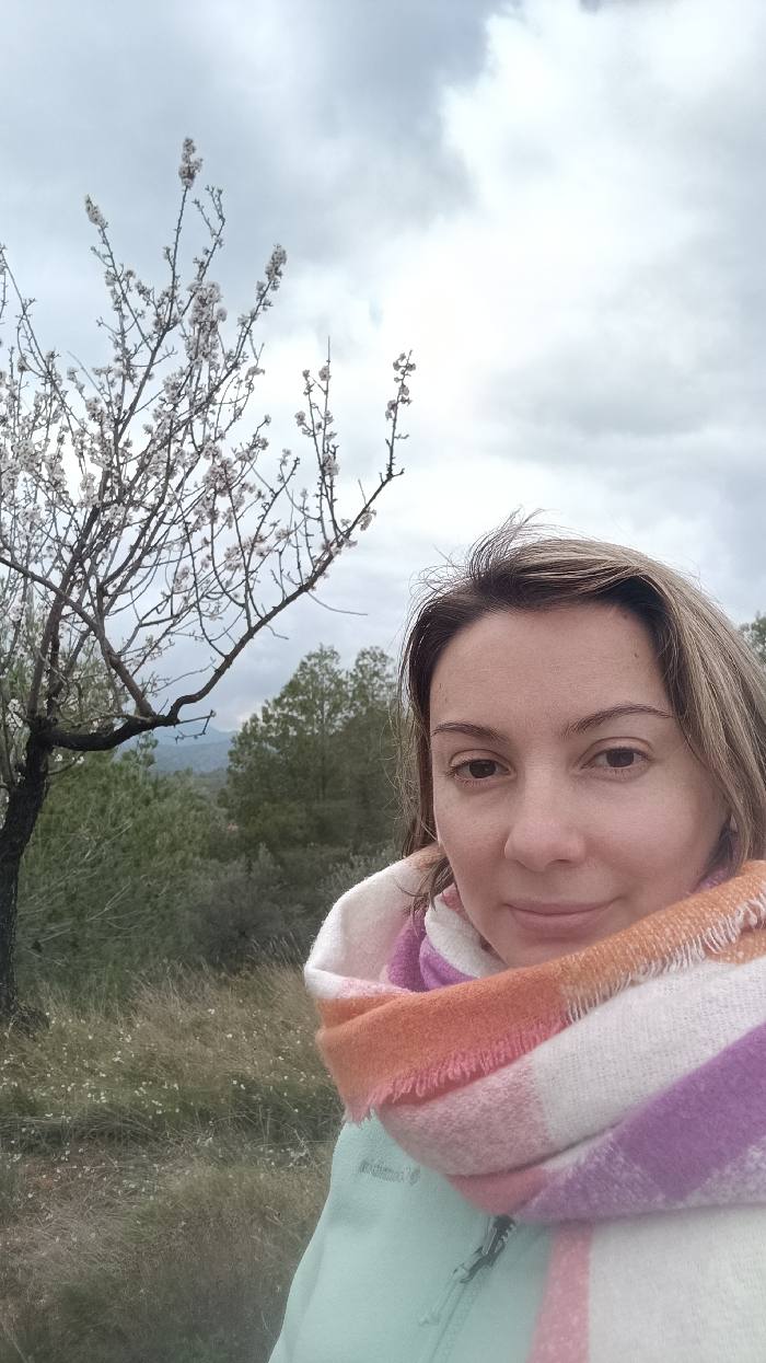 A close-up portrait depicts an adult female positioned outdoors within the city of Jérica, Spain. The subject, a Caucasian woman with light brown hair, occupies the right half of the frame, looking directly into the camera with a subtle smile. She wears a light green zippered jacket and a thick, multi-colored striped scarf featuring shades of white, pink, orange, and purple. To the left, a flowering tree with white blossoms on bare branches is visible, extending into the upper left quadrant. The immediate ground consists of tall, dry grass interspersed with small white flowers. The background features a dense expanse of green trees, likely a forested area, with distant hills or mountains under a heavily overcast, light grey sky.