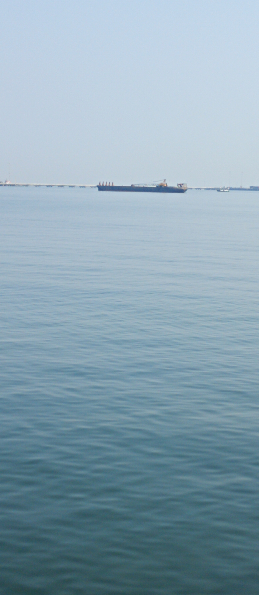 The image depicts a wide expanse of calm blue water, with subtle ripples disturbing the otherwise smooth surface. In the distance, a long pier stretches across the horizon, supported by numerous pilings. A large cargo ship, dark in color and rectangular in shape, is docked or anchored near the pier, its silhouette distinct against the pale sky. The sky itself is a pale, hazy blue, devoid of clouds, suggesting a clear day. The scene appears to be a maritime or port area, likely in a coastal city. Given the specific location of Probolinggo, Indonesia, this image likely captures a view of the Probolinggo Port, a significant shipping hub on the northern coast of East Java, known for its cargo and passenger services.