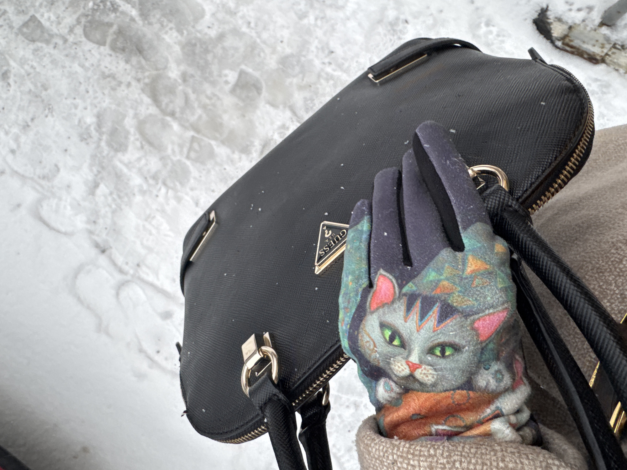The image captures a close-up, high-angle view of a black handbag and a hand wearing a distinctive glove against a snow-covered background, likely in Perm, Russia. The main subjects are a textured black handbag with gold-colored hardware and a gloved hand.
The black handbag, possibly made of faux leather, features a metallic triangular logo with the text "GUESS U.S.A." visible on it. It has gold-toned zippers and rectangular buckle details. Small flakes of snow are visible on its surface, indicating an outdoor winter setting.
The hand is adorned with a colorful glove featuring a stylized grey cat with vibrant green eyes, pink ears, and an orange-red scarf or collar. The cat's head is surrounded by a pattern of colorful geometric shapes. Part of a beige, knitted or textured fabric, possibly a coat or sleeve, is visible below the glove.
The background consists of white, uneven snow, suggesting footprints or an undisturbed layer. The overall scene depicts a person holding their bag and wearing a unique glove in a snowy environment. No specific activity beyond holding the items is clearly discernible. The time of day appears to be daylight due to the bright white snow.