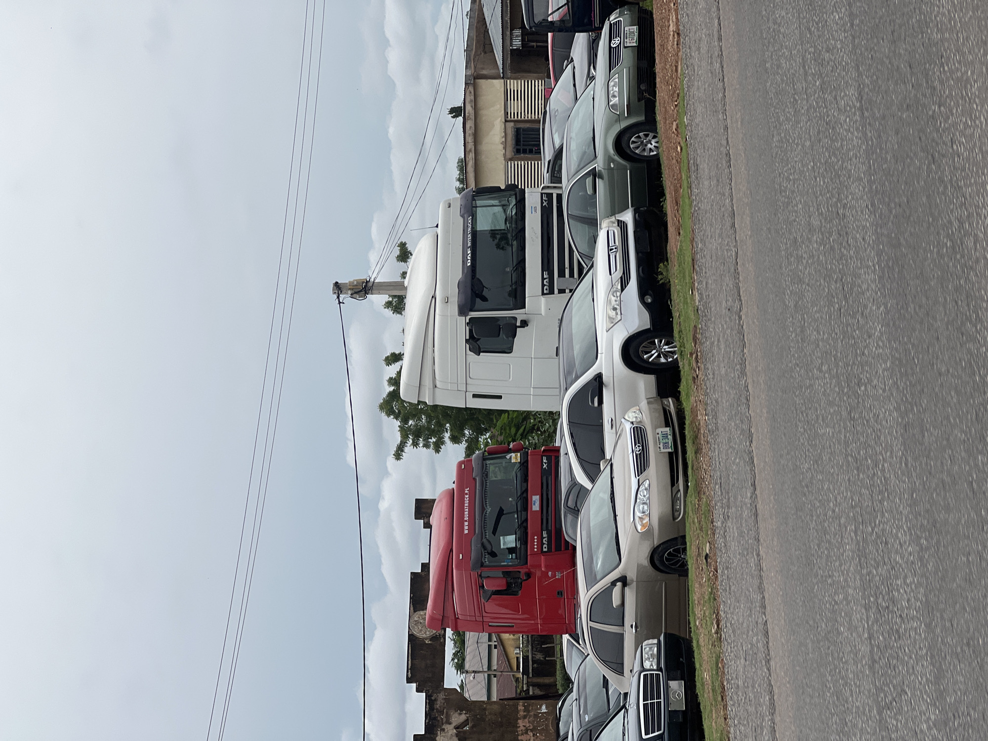 The image shows a street scene in Jalingo, Nigeria, featuring a variety of vehicles parked on what appears to be a lot. Prominently displayed are two large DAF trucks, one red and one white, positioned behind several passenger cars. The red truck has "www.runatruck.pl" written on its side. The passenger cars include a gold sedan, a silver sedan, and a black sedan. The environment is overcast with a cloudy sky. In the background, there are buildings and trees. The overall impression is of a vehicle sales lot or storage area.