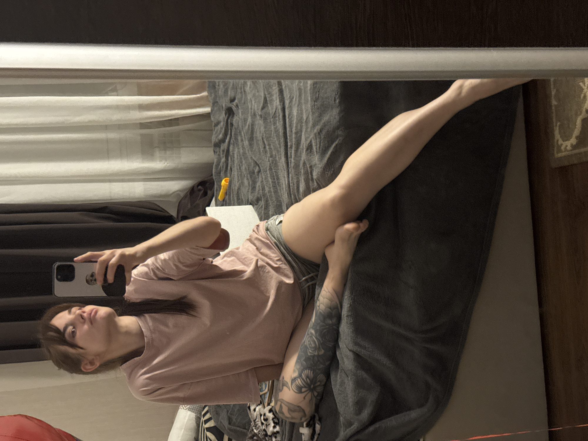 A woman with brown hair lies on her back on a gray blanket on a bed, taking a selfie in a mirror. She is wearing a pink t-shirt and gray shorts. Her left arm is tattooed. The room appears to be a bedroom. The location is not discernible from the image.