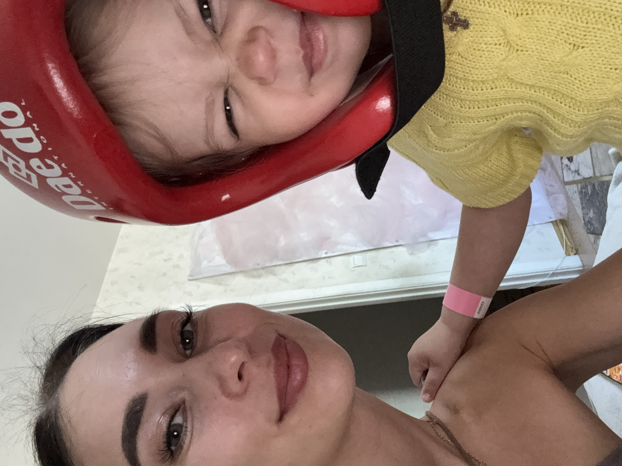 This vertical, close-up image captures an intimate indoor moment between an adult woman and a baby or toddler, likely a selfie taken in Chaikovsky, Russia.
In the foreground, the adult woman is positioned lower in the frame, with her head slightly tilted. She has dark hair, prominent eyebrows, and is looking directly at the camera with a neutral to softly smiling expression. Her left eye, nose, and lips are clearly visible. She wears a thin, light-colored chain necklace around her neck, and her shoulder and upper chest are visible, suggesting a sleeveless or off-shoulder top.
Above and slightly behind her, a baby or toddler is featured, wearing a bright red protective headgear that covers much of their face. White text "Dae Do" and "International" is clearly visible on the headgear, indicating it is likely martial arts equipment, possibly for Taekwondo. Despite the headgear, the child's wide, dark eyes and a playful, open-mouthed grin are discernible, conveying a happy or excited emotion. The child is dressed in a yellow knitted top with a textured pattern and has a pink wristband on their left arm, the text on which is illegible. One of the child's hands rests gently on the adult's shoulder or chest area.
The background is simple and somewhat out of focus, consisting of a light-colored wall or surface and some indistinct patterns, possibly suggesting tiled surfaces or a window/door frame. The lighting appears to be natural daytime light. The image conveys a warm, cheerful, and connected interaction between the two subjects.