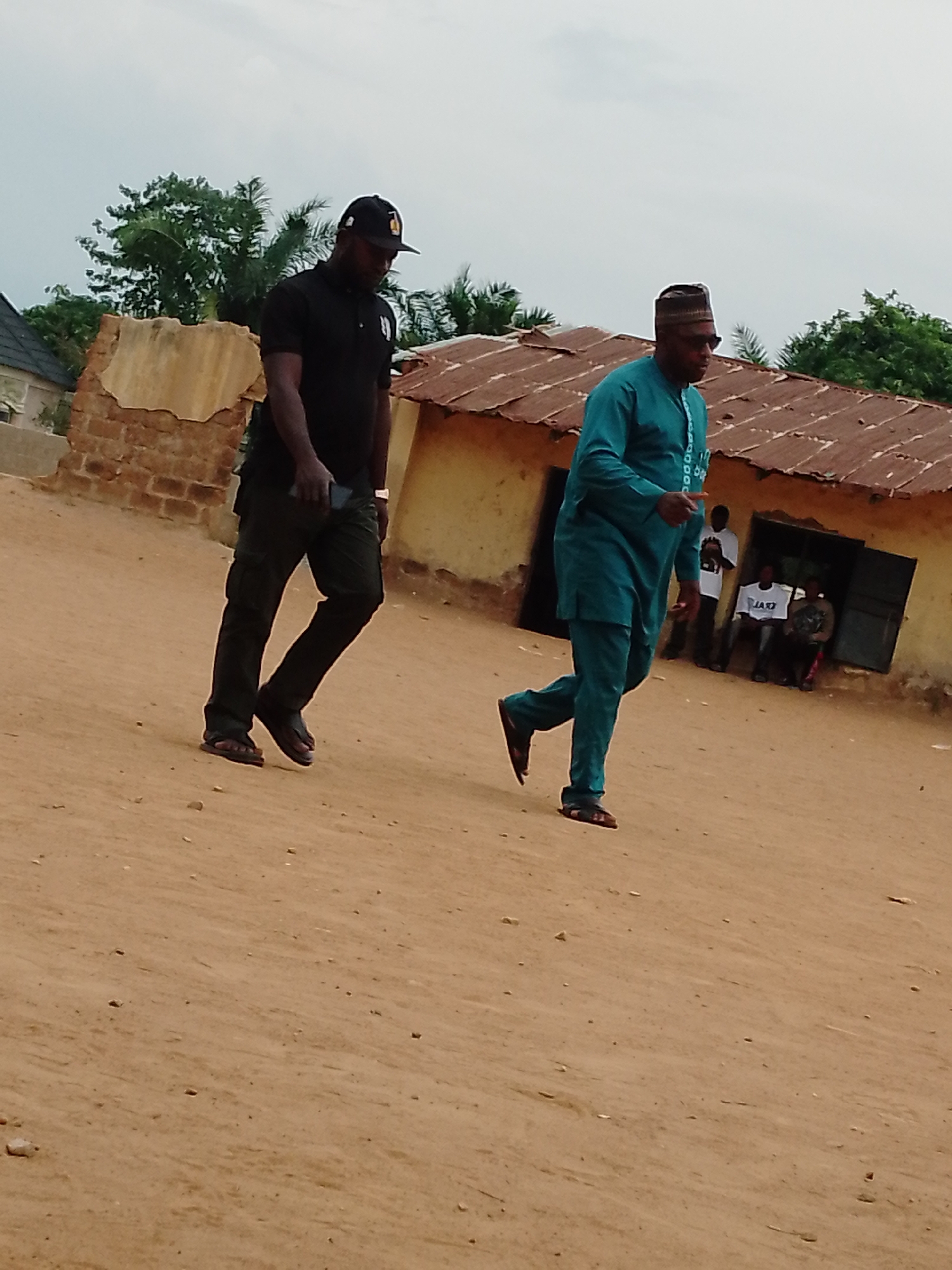 Two men are walking on a dirt road in front of a run-down building in an outdoor location in the city of Unknown, Unknown. The man on the left is wearing a black baseball cap, black polo shirt, dark trousers, and sandals. He is holding a dark object, possibly a phone, in his left hand. The man on the right is wearing a blue traditional outfit with a matching cap and sandals. He is also wearing sunglasses. Behind them, in front of the building, there are three people sitting, two wearing white shirts with text. A white sign with black text is also visible in the background. The buildings are dilapidated with brown walls and rusty roofs. Trees are visible in the background under an overcast sky.