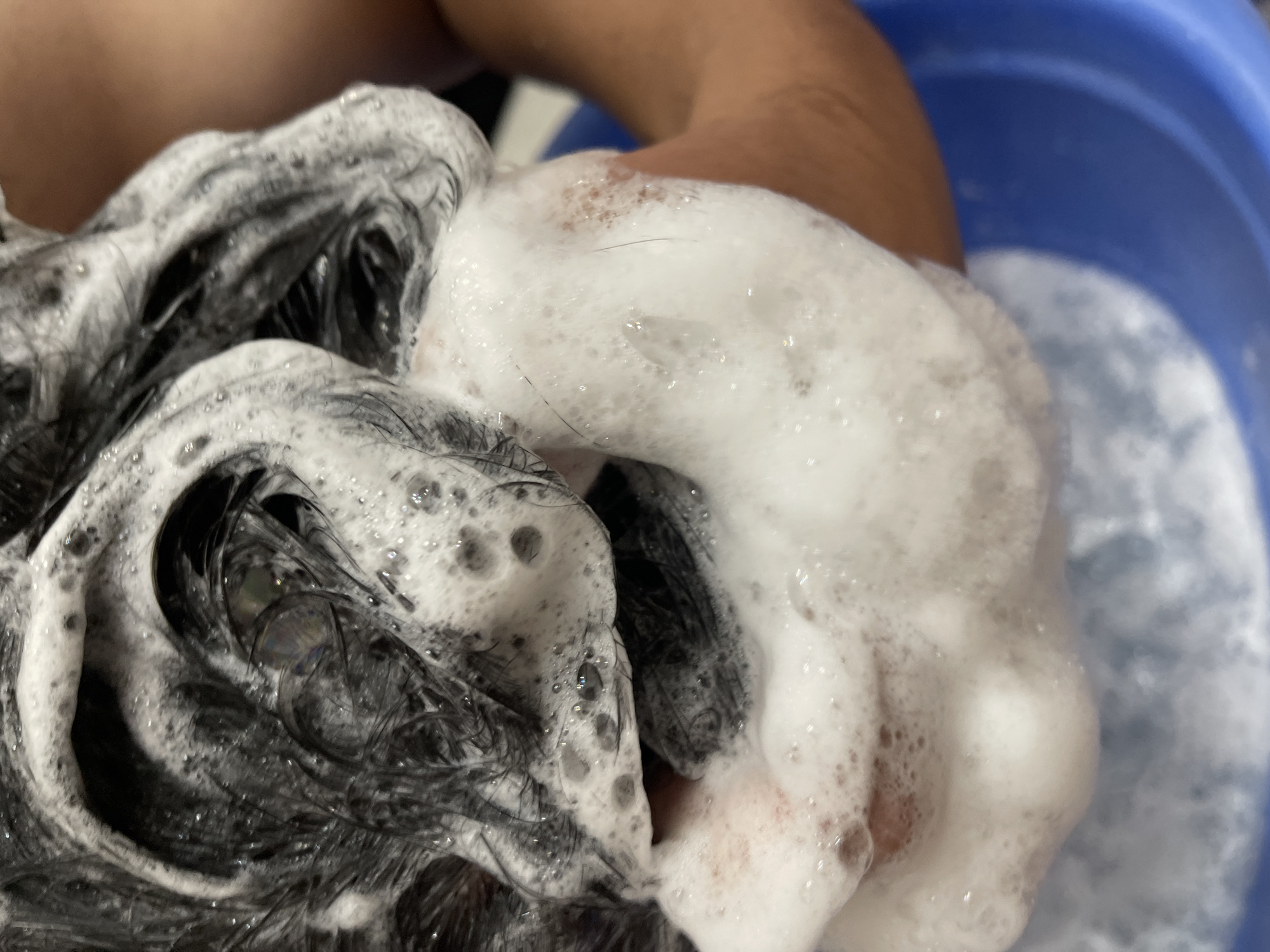 In a close-up, overhead shot, a person's dark hair, slick with white shampoo lather, is being washed. Bubbles cling to individual strands and form soft mounds. A hand, with light brown skin, is visible, cupping some of the soapy hair and obscuring a portion of the scalp. The background shows a blurred blue basin, suggesting the action is taking place in a bathroom or wash area. There are no identifiable landmarks or clues to pinpoint the location within Bishkek City, Kyrgyzstan.