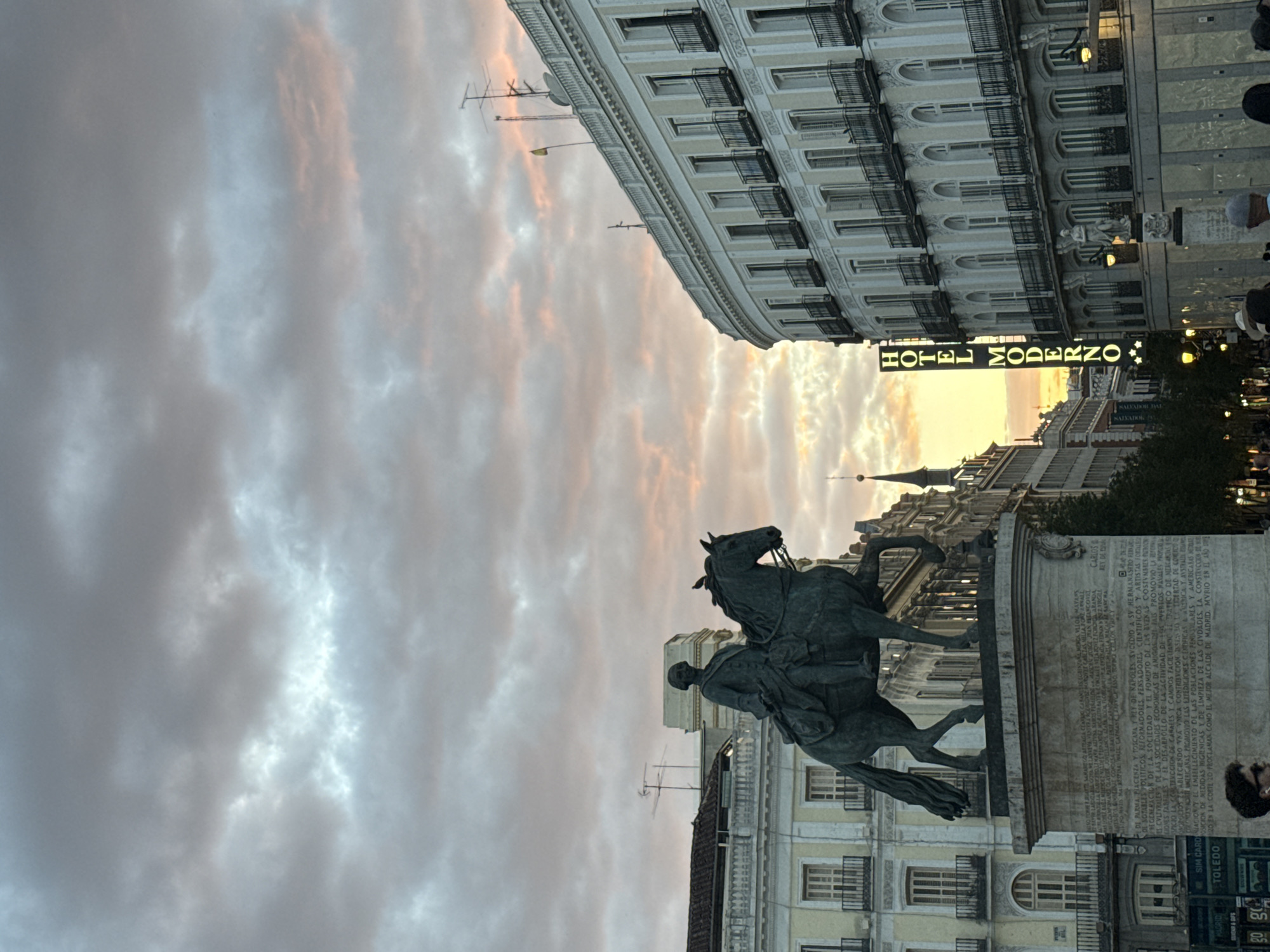 An equestrian statue stands prominently in the foreground of a bustling city square or street scene at sunset or twilight. The image is taken from a low angle, looking upwards, emphasizing the grandeur of the statue and the surrounding architecture against a dramatic sky.
The central subject is a dark bronze-colored equestrian statue, depicting a historical figure on horseback, with the horse in a rearing posture. The statue rests on a large stone pedestal, which is covered in extensive Spanish text. Key phrases deciphered from the text identify the figure as "CARLOS III REY DE ESPAÑA" (Charles III King of Spain), mentioning his reign, various political and cultural reforms, promotion of trade and infrastructure, scientific expeditions, and his reputation as the "MEJOR ALCALDE DE MADRID" (best mayor of Madrid), along with his death year, "MDCCLXXXVIII" (1788).
Behind the statue, a grand, multi-story building with ornate facades, balconies, and many windows dominates the mid-ground. A prominent vertical sign illuminated in yellow on this building clearly reads "HOTEL MODERNO". Other classical European architecture is visible further down the street. The sky above is filled with dynamic clouds, tinted with hues of orange, pink, and grey from the setting sun, with brighter patches of sunlight breaking through. Several antennas and satellite dishes are visible on the rooftops of the surrounding buildings.
At the very bottom right of the image, the tops of people's heads are visible, suggesting a busy pedestrian area. Streetlights are beginning to illuminate, adding to the evening atmosphere. Other smaller signs with text like "SIM CARD" and "TOLEDO" can be seen near street level. The architectural style, the specific hotel, and the detailed inscription on the statue confirm the location as Madrid, Spain, likely near Puerta del Sol.
