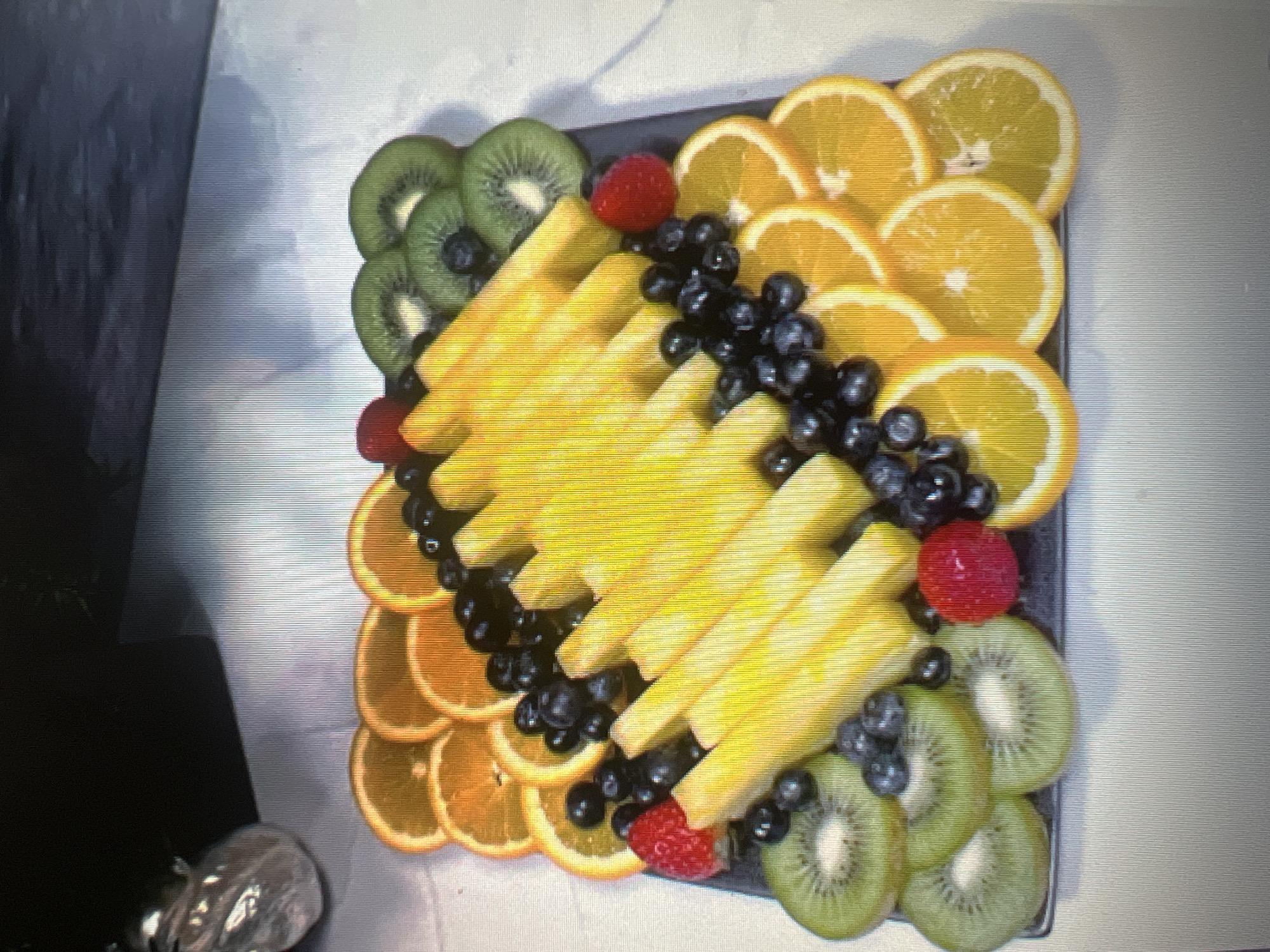 A fruit platter is displayed on a dark square platter. The platter contains slices of oranges, kiwi slices, blueberries, raspberries, and pineapple sticks arranged in a decorative pattern. The platter is placed on a white surface.
