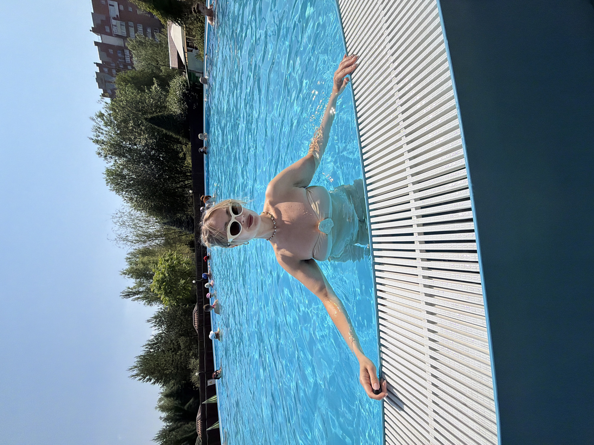 A young woman with blonde hair and white-rimmed sunglasses is partially submerged in a bright blue swimming pool. She is wearing a light-colored bikini and a beaded necklace. Her right arm is extended, resting on the edge of the pool, which has a white, slatted surface. The water is clear and reflects the sunlight, creating ripples.
In the background, several other people can be seen in the pool, some wearing swim caps. Beyond the pool, there are lush green trees and a tall, multi-story brick building under a clear blue sky. The presence of the building and trees suggests an outdoor urban or suburban setting. The sunlight indicates it is daytime, likely on a warm, clear day. The overall mood appears relaxed and recreational, consistent with a day at a swimming pool. The location context provided is Krasnoyarsk, Russia. No visible text is present in the image.