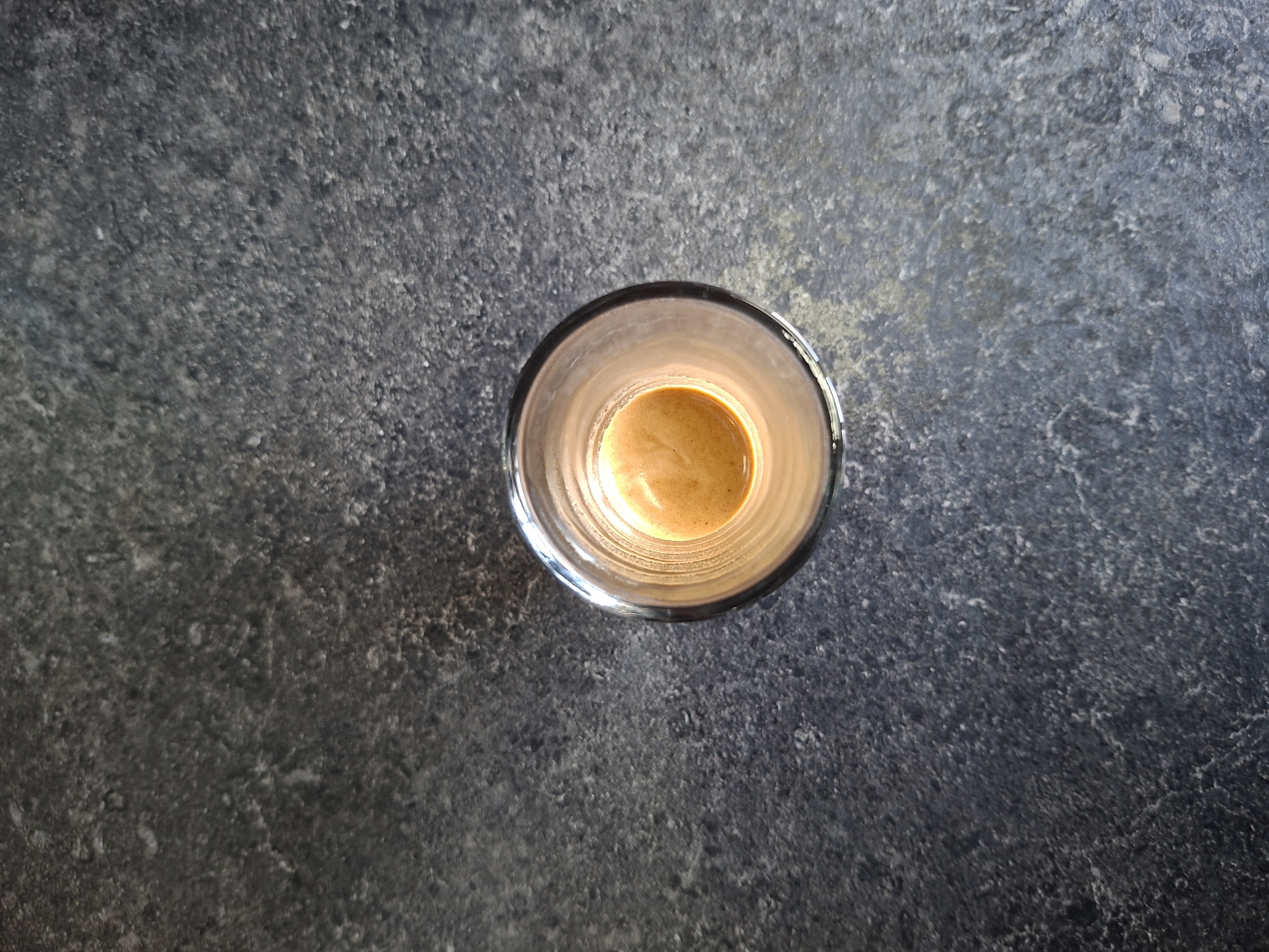A top-down, medium shot shows a single shot of espresso in a clear glass on a dark, textured surface, possibly a granite countertop. The espresso has a light brown crema on top. The surface is speckled with lighter and darker gray tones, giving it a mottled appearance. The lighting suggests an indoor setting, with a warm light reflecting off the crema and the rim of the glass. The image is a still life with no people or activity depicted. The location in Chișinău, Moldova, is not determinable from the visual information provided.