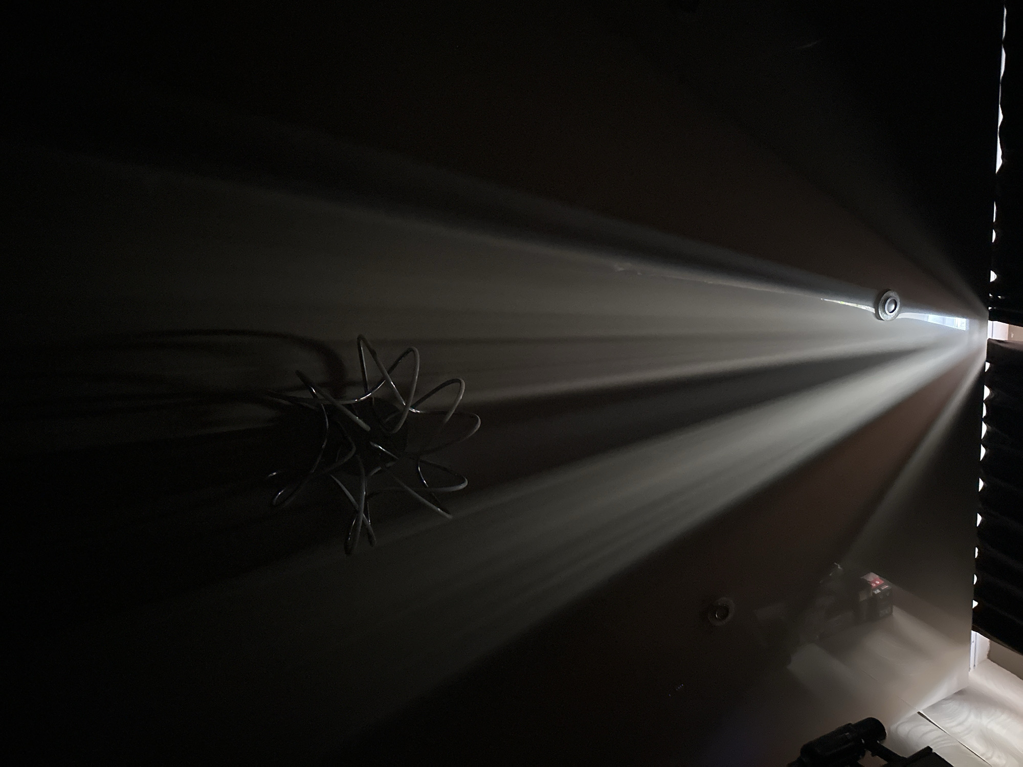 The image shows light streaming into a darkened room through a set of blinds. The light illuminates a wall with a wire sculpture and the lower part of the room with visible objects on the floor. Location is unknown.