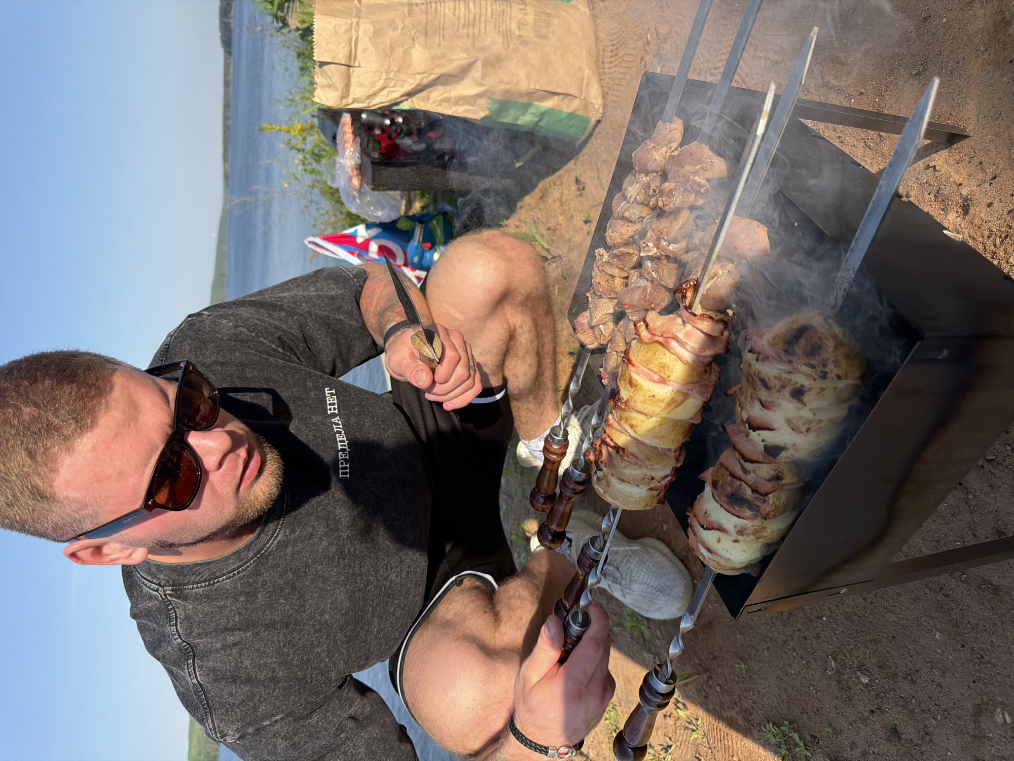A man is grilling meat on a skewer over a portable barbecue grill. He is holding a knife in one hand. The man is wearing sunglasses, a gray shirt, and black shorts. The grill contains three skewers of meat, one on the right, one in the middle, and one on the left. The right skewer has cut pieces of meat. The middle and left skewers both contain a cylindrical piece of white food wrapped with strips of bacon. Smoke rises from the grill. The location is outdoors on dirt. In the background is a body of water, plants and a paper bag. The text on the man's shirt is vertically aligned, starting with a letter which looks like a 'P' and continues with the letters "REДELA HET". It is unknown if this is the city of Chaykovsky, Russia.