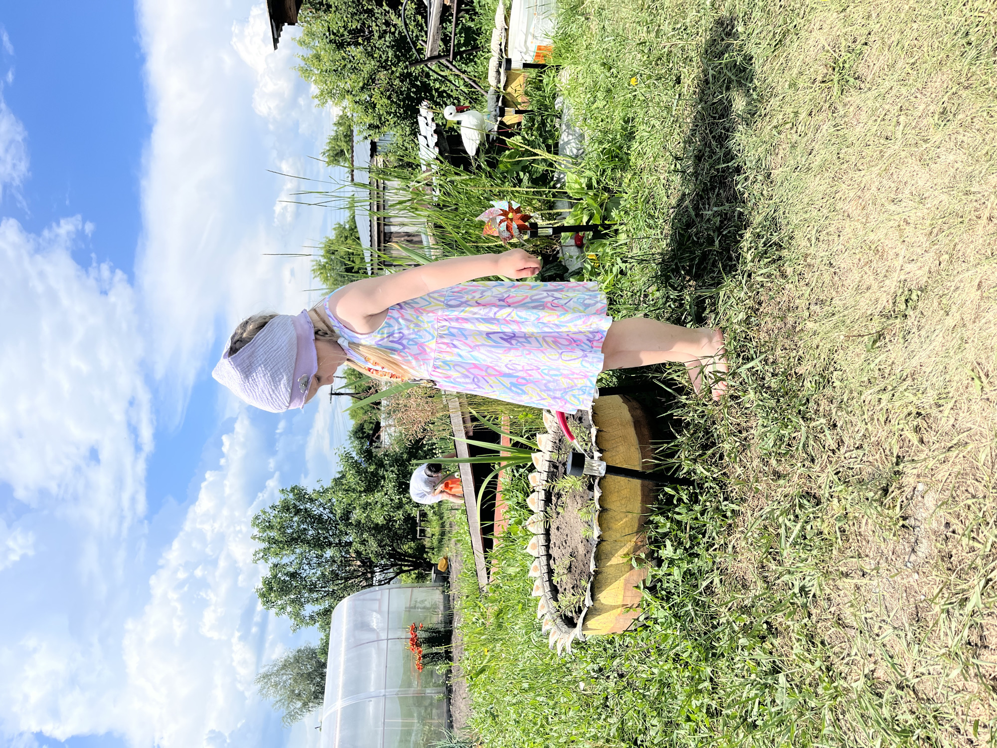 In a rural outdoor setting, a young girl with blonde hair, wearing a light purple headscarf and a colorful sleeveless dress, stands in a grassy yard. She is looking down and to her right. To her left, a raised circular garden bed, possibly made from a repurposed tire, contains soil and small green plants. A small solar-powered garden light is positioned in front of the bed. Behind the girl, a greenhouse with orange flowers is visible, along with a person in a white shirt tending to something orange. Further back, there are trees and buildings. A white decorative bird statue stands near some vegetation and a small, colorful pinwheel is stuck in the ground. The sky above is blue with scattered white clouds. The overall scene suggests a sunny day in a garden or rural property, likely in the village of Sukhrinskoye, Russia, given the provided context.