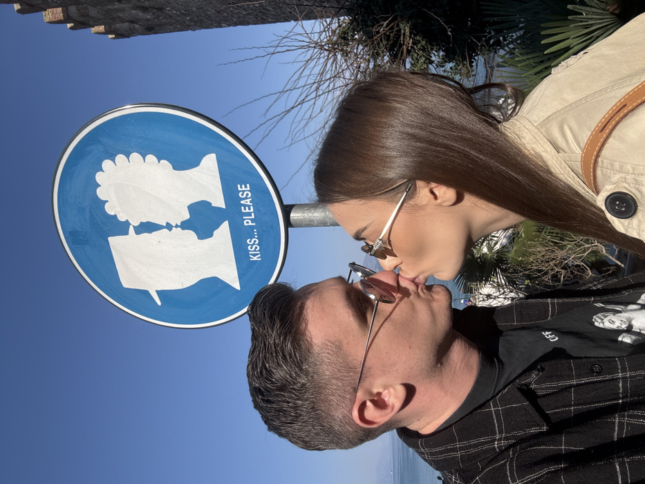 A man and a woman are shown kissing directly beneath a prominent blue circular sign that reads "KISS... PLEASE" in Sirmione, Italy. The sign, mounted on a pole, features white silhouettes of a man in a fedora and a woman with curly hair facing each other, positioned to appear as if kissing.
The man, on the left, has short, dark hair styled to the side and wears silver-rimmed round sunglasses with a light tint. He is dressed in a dark plaid overshirt over a black t-shirt with a white graphic print. The woman, on the right, has long, straight dark brown hair and wears gold-framed sunglasses. She is wearing a beige or light brown trench coat over her left shoulder, with a brown strap from a bag visible. Both individuals have their lips puckered in a kiss, with the man's hands not visible and the woman's right arm, if any, obscured by the frame.
In the background to the left, a body of water, likely Lake Garda, is visible under a clear blue sky. Mountains are faintly visible across the water in the distance. To the right, a section of the Scaliger Castle walls, characterized by crenellated battlements, is visible, indicating the location is near the castle's entrance. Sparse, leafless tree branches and green foliage are also present in the mid-ground, suggesting an outdoor, possibly early spring, setting. The scene is brightly lit by natural sunlight.