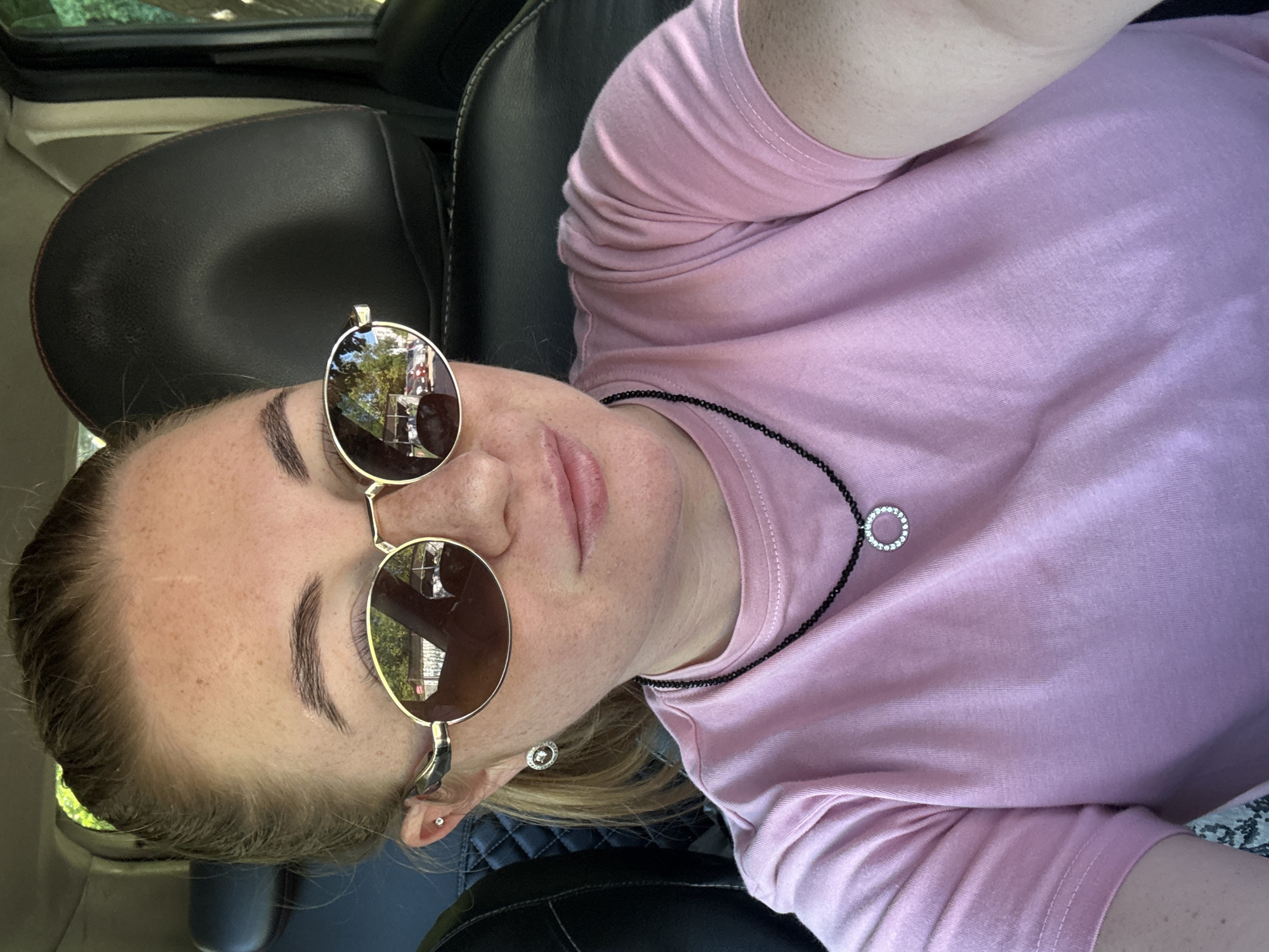 A medium shot captures a young white woman with blonde hair pulled back in a ponytail, wearing large, gold-rimmed sunglasses and a pink t-shirt, as she sits in the driver's seat of a car. She is wearing a black beaded necklace with a circular pendant adorned with small, clear stones, and matching earrings. Her reflection in the sunglasses shows trees and buildings. The interior of the car is visible, with dark leather seats. The scene appears to be outdoors during the day, with sunlight casting shadows and reflections. The woman is looking directly at the camera.