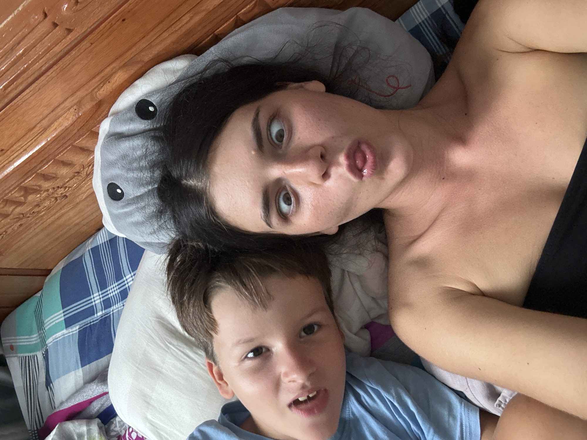 A boy and a woman are lying in bed, looking at the camera. The boy is on the left side of the frame, wearing a blue shirt. He has brown hair and is looking directly at the camera with his mouth slightly open. The woman is on the right side of the frame, wearing a strapless black top. She has dark hair and is making a funny face with her eyes crossed and her tongue sticking out. She is leaning on a grey stuffed animal with large black eyes. The bed is covered with a blue and white checkered blanket and a white pillow. The background appears to be a wooden headboard of the bed. The image appears to have been taken indoors.
