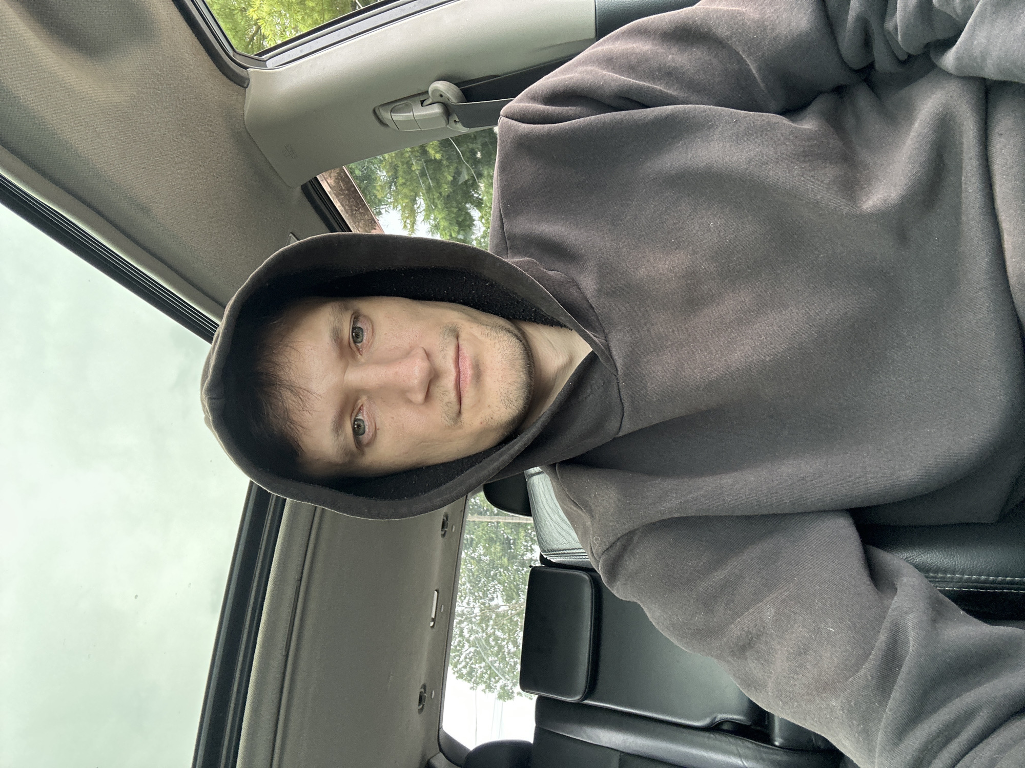 A man in a dark grey hooded sweatshirt is sitting in the driver's seat of a car. The sunroof of the car is open, revealing a cloudy sky. The interior of the car is visible, including the black leather seats in the back. Outside the car, trees and foliage can be seen. The image is a selfie, taken from a slightly low angle, looking up at the man's face. The man has light skin, short brown hair, and light green eyes. He is looking directly at the camera with a neutral expression. There are no other people or objects in the immediate vicinity of the man. The scene suggests a casual, everyday setting, possibly a person taking a break or on a journey. The location is not specified in the image itself, but the question asks to include the city of Makiv, Ukraine, which is extraneous information not derivable from the visual data.