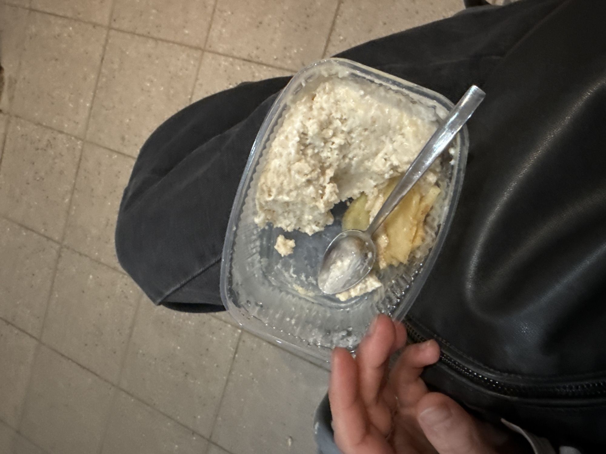 An overhead shot captures a person's hand interacting with a clear plastic container of food, placed on or near dark fabric, with a tiled floor visible in the background.
The central subject is a rectangular, transparent plastic food container, appearing to be a disposable takeout or ready-meal container. Inside, there is a substantial amount of a light-colored, lumpy, creamy food, possibly oatmeal, cottage cheese, or a similar textured dish. Mixed within this are several yellow or golden pieces, which could be fruit slices like apple or pear. A silver-colored metal spoon rests in the food, suggesting it is being or has been eaten.
To the right and underneath the container, a dark-colored fabric is visible, which could be part of a jacket, bag, or clothing item, possibly made of leather or a similar smooth material. A black zipper is clearly seen along the edge of this fabric. A person's right hand, with fingers slightly curled, is visible in the lower right corner of the frame, positioned near the container and the dark fabric.
The background consists of a light-colored, square-tiled floor with visible grout lines and a speckled texture, indicating an indoor setting. The overall lighting appears to be artificial. No text is clearly legible on any visible objects. The scene depicts a casual moment, likely a meal or snack.
