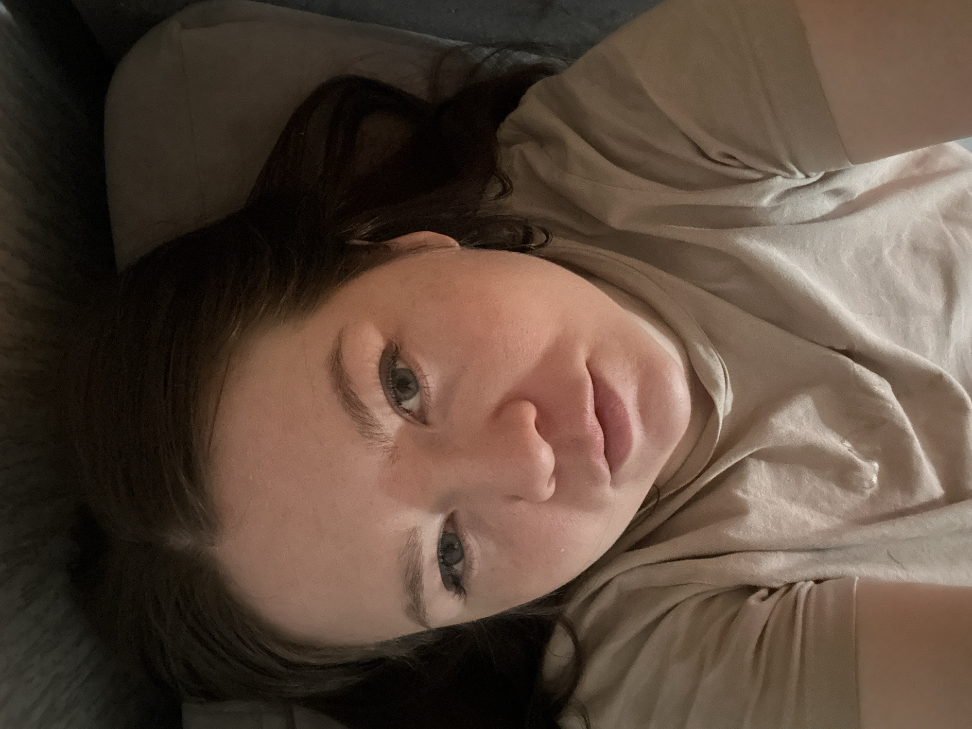 A woman with light skin and dark brown hair is lying down, looking directly at the camera. She has green eyes, a prominent nose, and full lips. She's wearing a light beige t-shirt, and her head rests on a gray or beige pillow or cushion. The background appears to be a couch or bed. The image is taken from a slightly elevated angle, looking down at the woman. There is no indication of this photo being in the city of Chaikovsky, Russia.