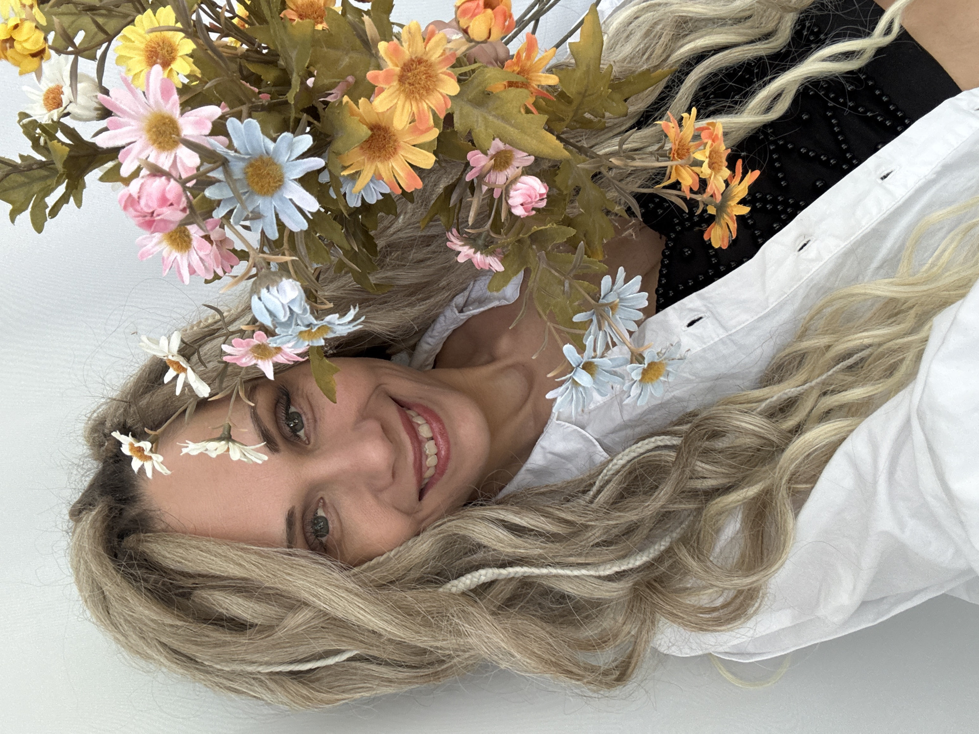 A woman with blonde hair and a flower crown lies on a white surface. She wears a white shirt with a black top underneath. Her hair is styled in loose waves with some strands braided. She is smiling and looking at the camera. She is holding a bunch of daisies in the colors of yellow, white, blue, pink, and orange. There are green leaves mixed in with the flowers. The image location is not determinable.