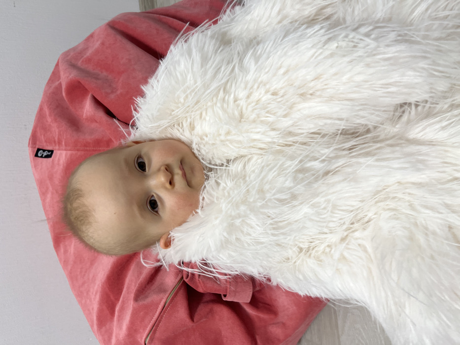 A baby lies nestled on a faded pink beanbag. The child is mostly enveloped in a fluffy, white blanket that frames their face. The baby has fair skin, a few freckles, and brown eyes, looking directly at the camera. The beanbag has a tag with "GP" stitched on it. The background is plain, suggesting an indoor setting with a white wall and a light-colored floor.