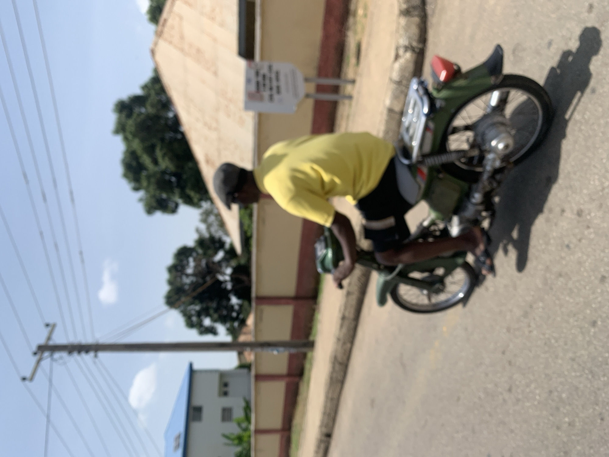 A person wearing a yellow t-shirt, black shorts, a cap, and sandals is riding a green motorcycle. The motorcycle has a rack on the back. The person is on a paved road, with a sidewalk and a wall to their left. Behind the wall, there are buildings and trees. A utility pole with wires is visible in the background. The scene appears to be in a city, possibly Jos, Nigeria, given the architectural style and vegetation. The sky is blue with some clouds.
