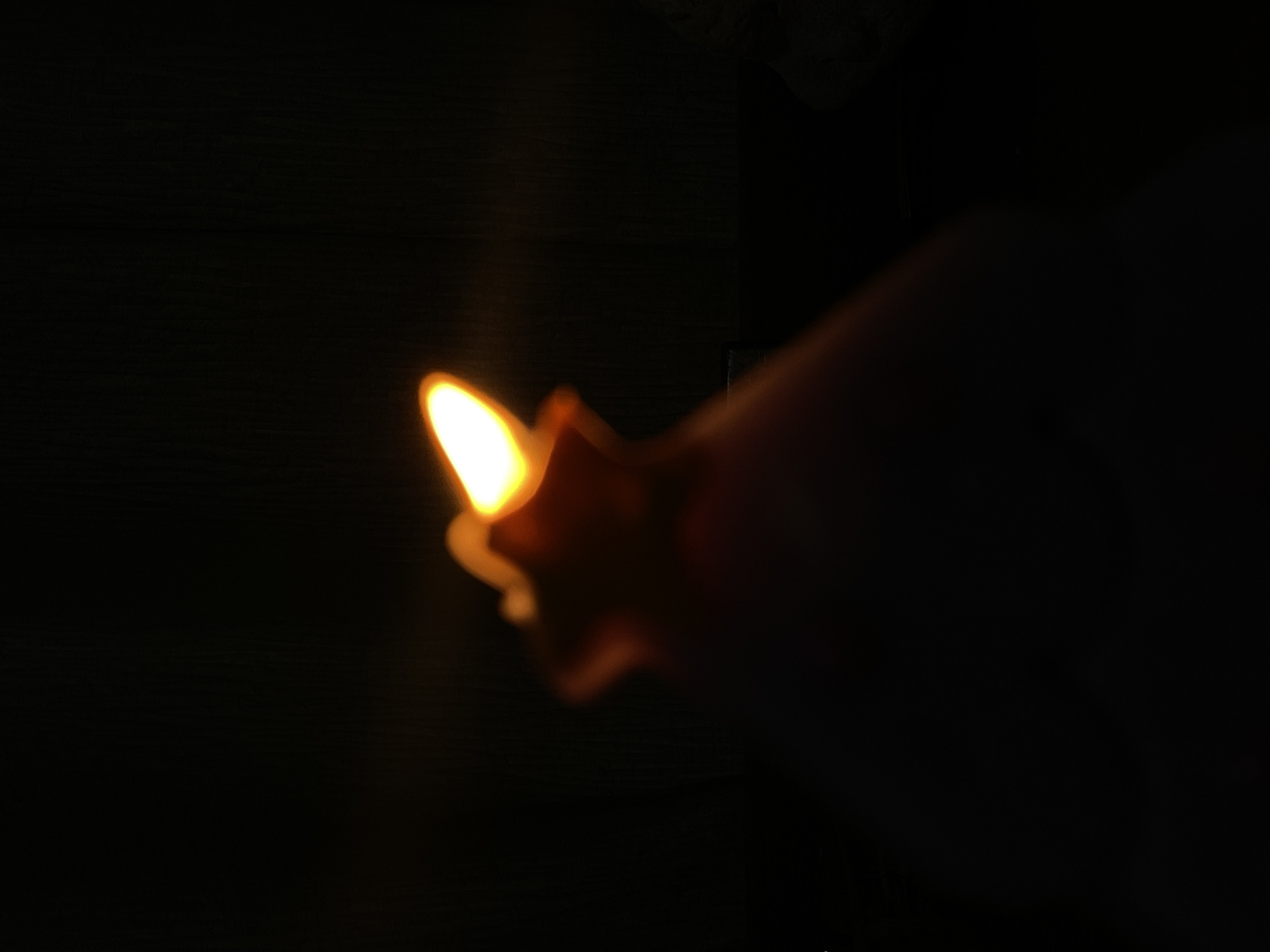The image captures a bright, warm light source set against a predominantly dark, out-of-focus background. The central subject is an intensely glowing, elongated light, with a bright white-yellow core that diffuses into an orange and amber glow around its edges. This light source has a distinct, somewhat pointed shape, resembling an artificial flame or an LED light rather than a natural candle flame.
Adjacent to and partially obscured by the light is a blurry, dark, reddish-brown shape, which appears to be holding or supporting the light source. This shape is indistinct due to significant blurring and low light, but its organic curves suggest it could be a hand or fingers.
The background is almost entirely black, indicating a very dark environment, likely night or a dimly lit indoor space. Faint, dark horizontal lines are barely discernible in some areas, hinting at a textured surface, possibly wood or a wall, but these details are heavily obscured by the darkness and blur. The overall scene is characterized by a strong contrast between the radiant light and the surrounding obscurity. There is no visible text in the image. The general impression is one of a small, isolated light cutting through profound darkness.