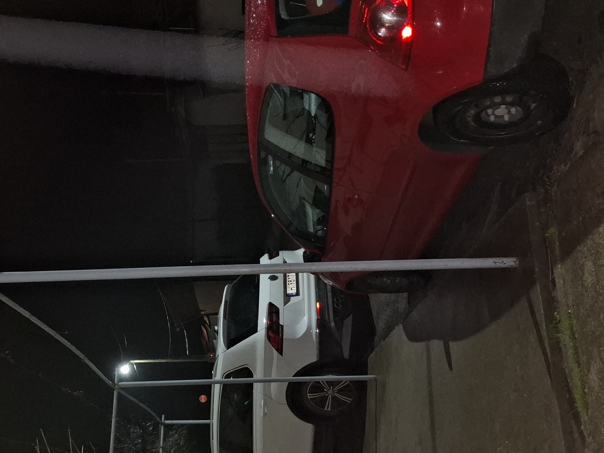 The image shows two cars parked on a street in Veliki Preslav, Bulgaria. The first car is red and positioned partially off the frame. The second car is white and is parked behind the red car. The license plate of the white car reads "H 9B17". A metal structure is also visible in the foreground. The environment appears to be a dimly lit area, possibly at night.
