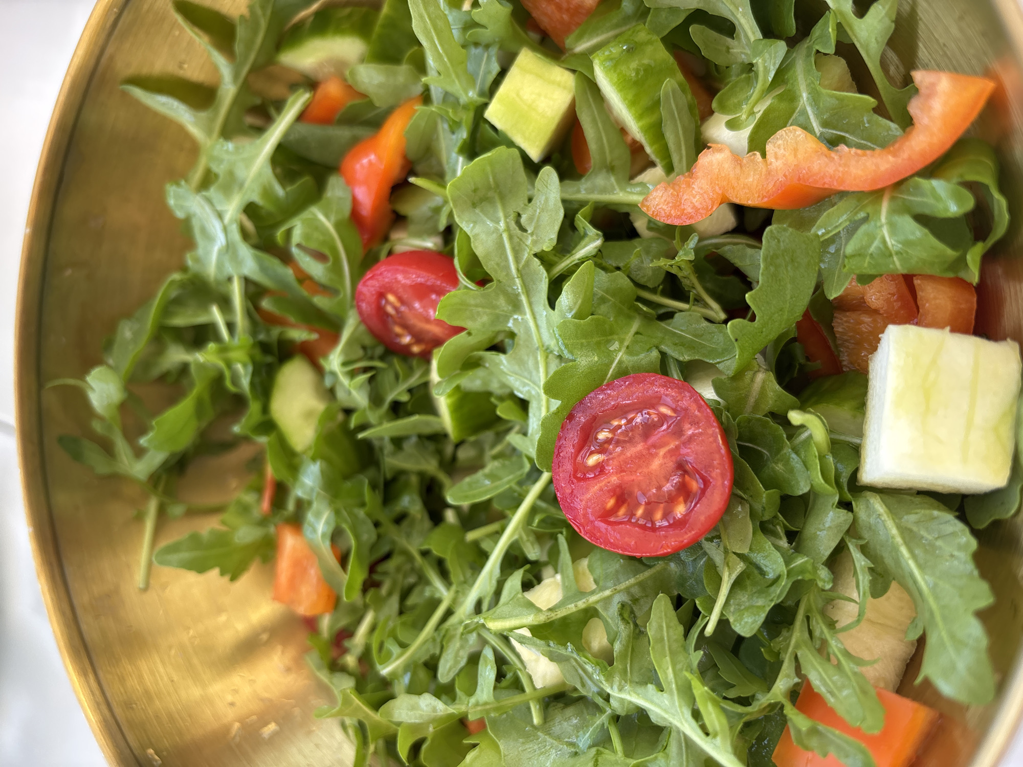 A close-up, eye-level shot captures a vibrant arugula salad served in a brass bowl. The salad is a medley of textures and colors, featuring bright green arugula leaves, diced cucumber, chopped red bell pepper, and halved cherry tomatoes. The tomatoes, in particular, are a focal point, their glossy red skins and juicy interiors vividly contrasting with the surrounding greens. The brass bowl, with its warm, metallic sheen, adds a touch of elegance to the rustic dish. The overall impression is one of fresh, healthy, and delicious food, ready to be enjoyed. The context provided is that this image was taken in the city of Razdolnoye, Russia.