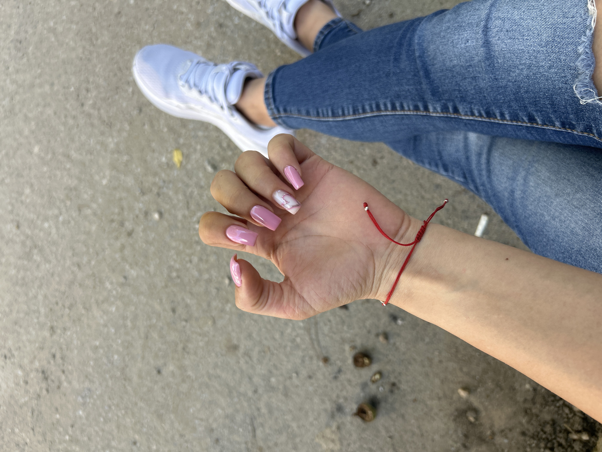 A person's hand with pink acrylic nails and a red string bracelet is shown. The nails on the index and ring fingers have a marble pattern with a heart shape. The person is wearing blue jeans and white sneakers. The background is a grey, textured surface.