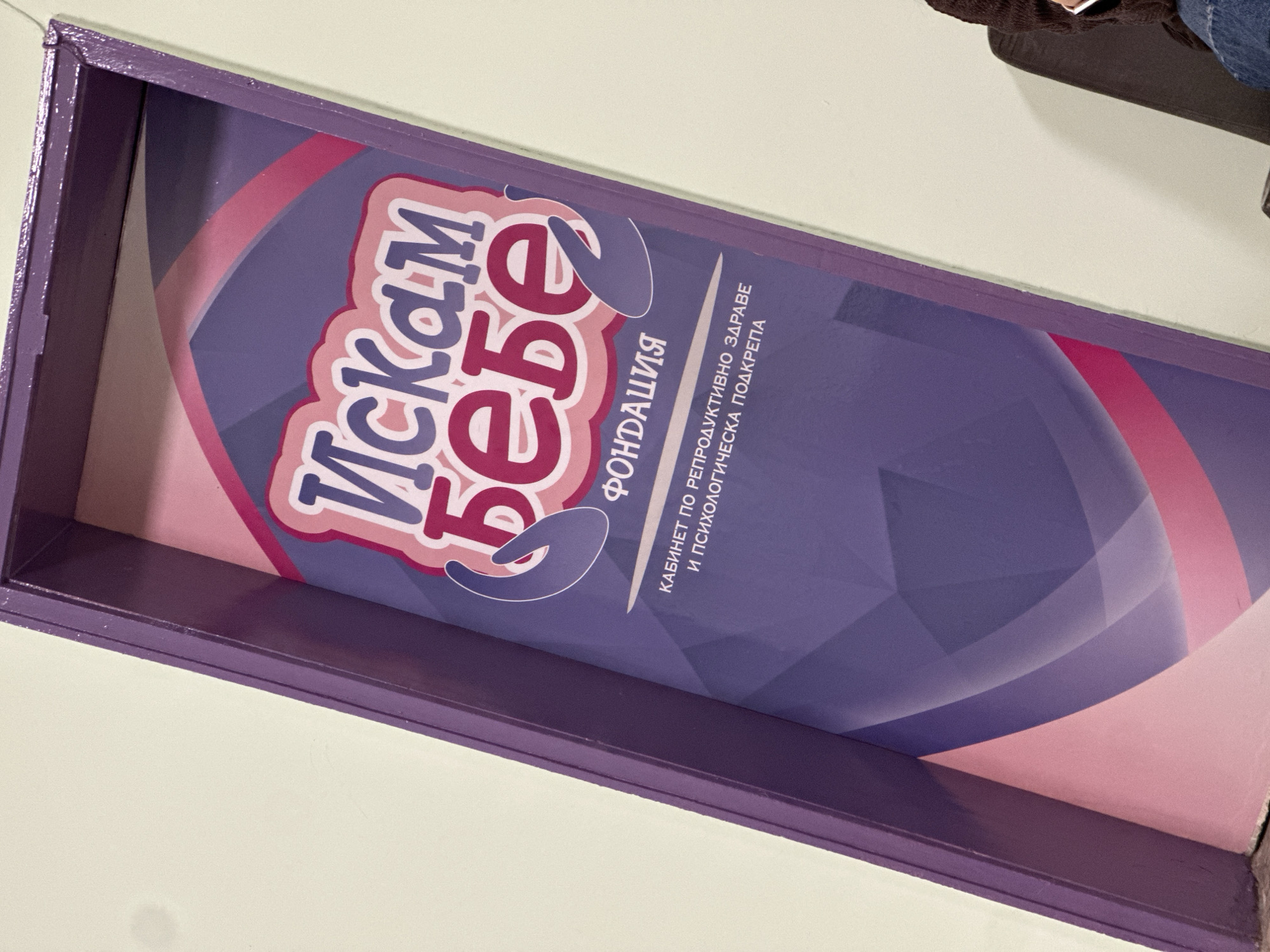 A close-up shot shows a purple-painted rectangular box or tray with an advertisement. The advertisement has a logo in Bulgarian Cyrillic reading "Искам бебе", which likely translates to "I want a baby". The text beneath the logo says "Фондация" (Foundation) and "кабинет по репродуктивно здраве и психологическа подкрепа" (office for reproductive health and psychological support). The logo and text are against a background of swirling pink and purple patterns. The box is sitting on a white surface. Part of someone sitting is visible, with brown cloth and jeans. There is no indication of being in Lovech, Bulgaria.