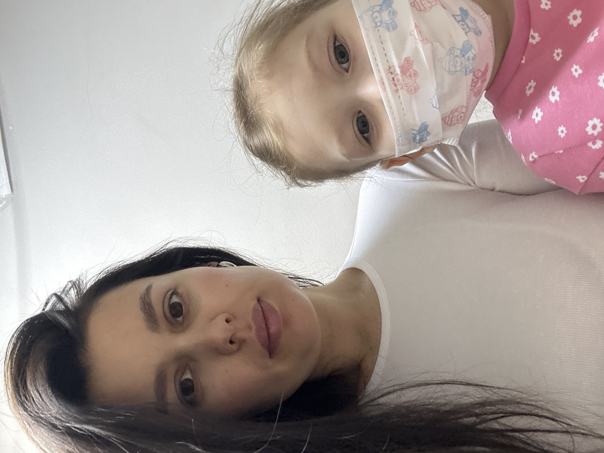 The image depicts a woman and a young child, both looking down at the camera. The woman has dark hair, brown eyes, and is wearing a white shirt. The child has light hair, is wearing a pink shirt with white flowers, and has a patterned face mask on. The background appears to be a plain white wall. The location is in the city of Chaikovsky, Russia.