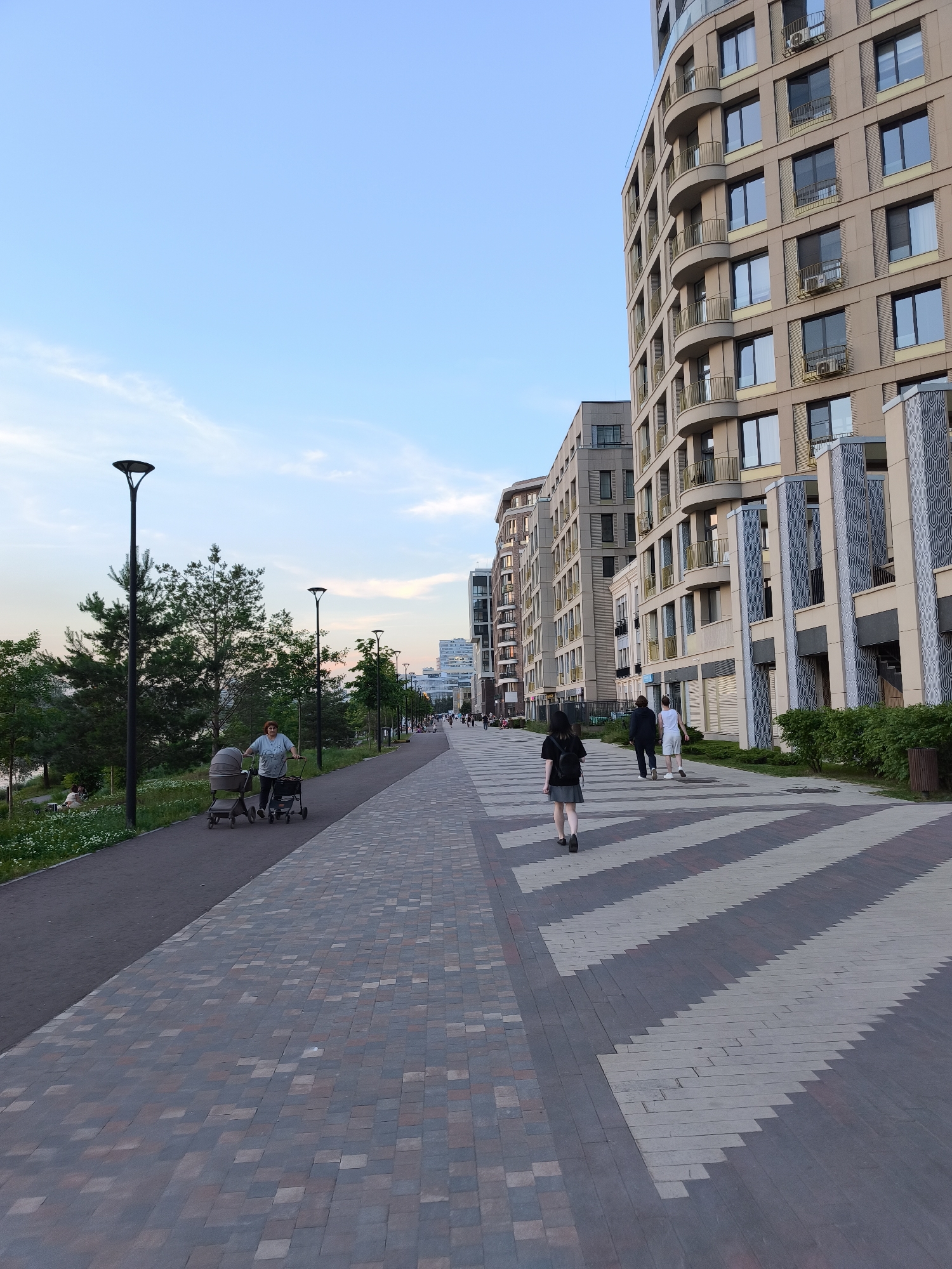 The image depicts a wide, paved pedestrian walkway in Moscow, Russia, featuring a modern apartment building on the right and trees and a park area on the left. The walkway is paved with a patterned mix of grey, brown, and reddish-brown interlocking pavers, with a zigzag pattern of light grey and dark grey pavers leading the eye into the distance. Several people are visible on the walkway.
In the foreground on the left, a woman is pushing a baby stroller and a walker. Further down the walkway, a young woman with dark hair and a backpack is walking away from the camera. Further still, a couple is walking away from the camera, the male figure wearing white shorts and a dark shirt, and the female figure in a dark shirt. In the distance, more people are scattered along the walkway.
The apartment building on the right is a multi-story structure with a curved facade, featuring numerous balconies with golden railings and large windows. The building's facade is a light beige color. Adjacent to the building, there are shops with closed storefronts.
To the left of the walkway, there is a grassy area with trees and a small body of water. The sky is a clear, pale blue with some wispy clouds, and the lighting suggests it is late afternoon or early evening. Streetlights are positioned along the walkway. In the far distance, more contemporary buildings can be seen, indicative of a developed urban area.