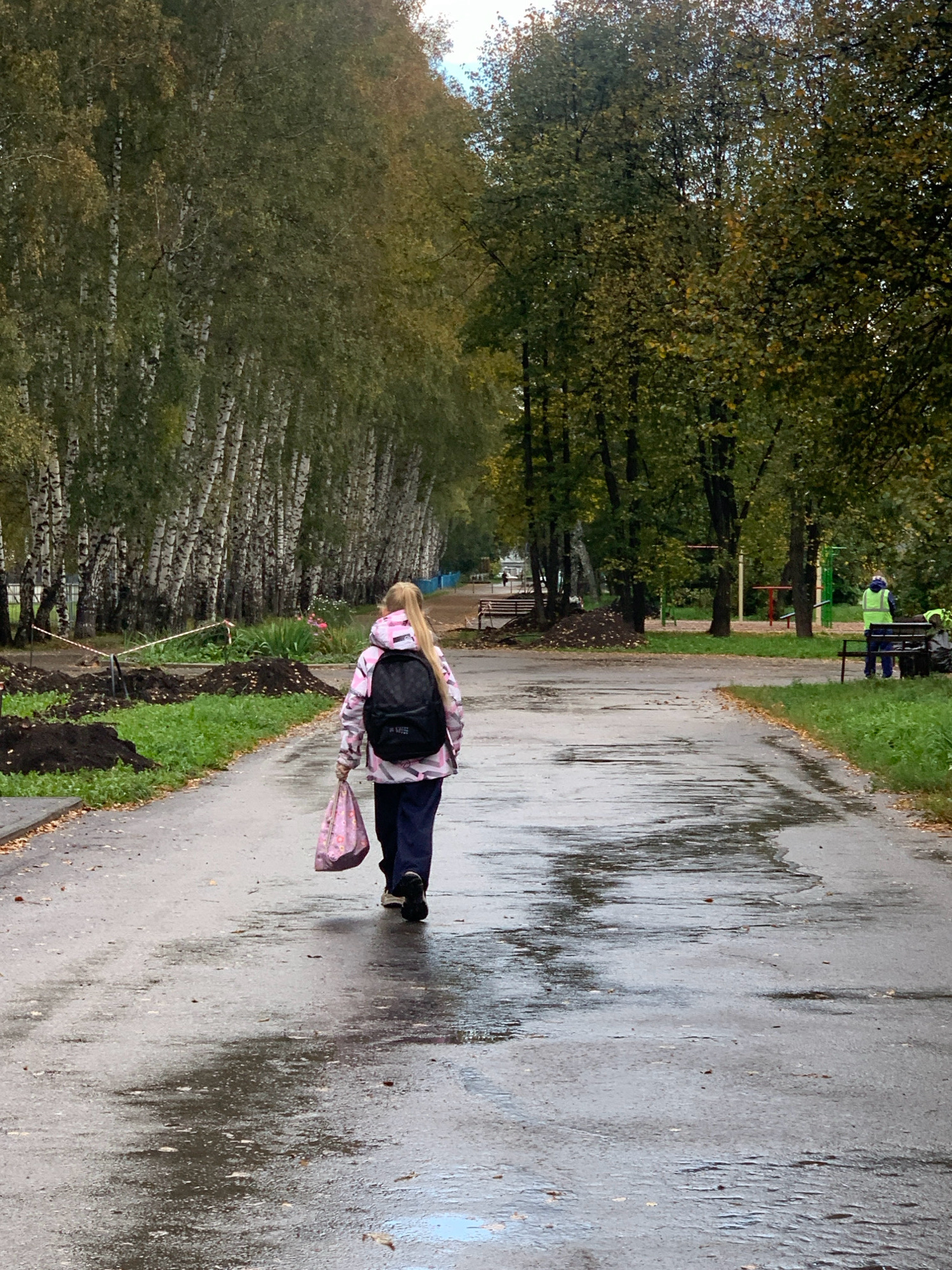 In Prokopyevsk, Russia, on an overcast day, a young person with blonde hair pulled into a ponytail walks away from the camera down a wet, paved path. The individual wears a pink and white patterned jacket, dark pants, and a black backpack. They are carrying a pink bag in their right hand. The path is flanked by trees, with a line of birch trees on the left and denser foliage on the right. Construction barriers and piles of dark soil are visible to the left of the path, indicating ongoing work. Further down the path on the right, two individuals in safety vests are present near a park bench, suggesting they might be park workers. The wet pavement reflects the surrounding environment, indicating recent rain.
