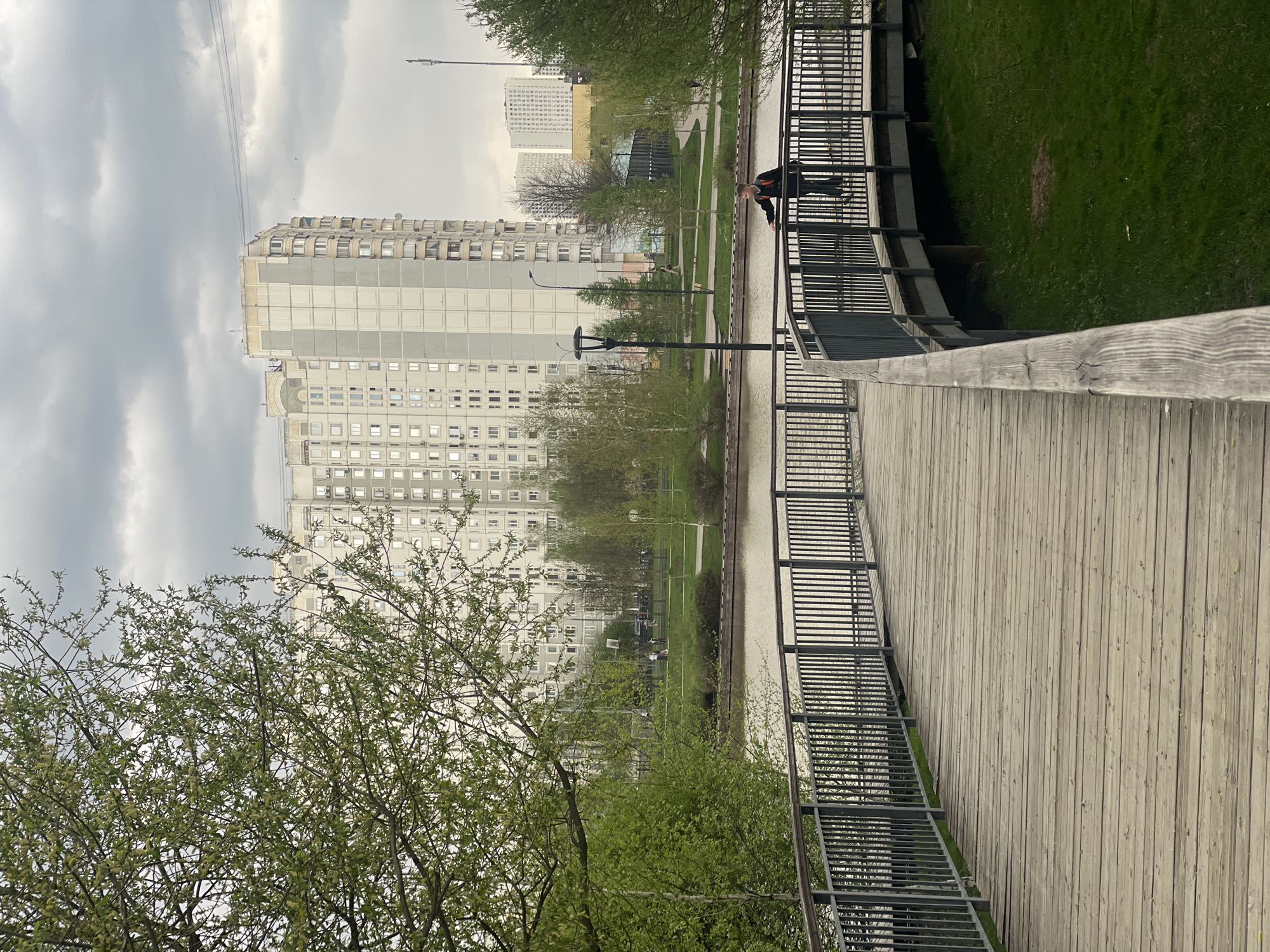 An eye-level perspective depicts a park in Moscow, Russia. A wooden bridge with metal railings dominates the foreground. A person stands on the bridge's far side, peering over the railing. Across the water, the park features green lawns and trees, with several people walking in the distance. Tall, grey, apartment buildings are visible in the background under a cloudy sky. A power line runs diagonally across the sky. A green leafy tree is in the left foreground.
