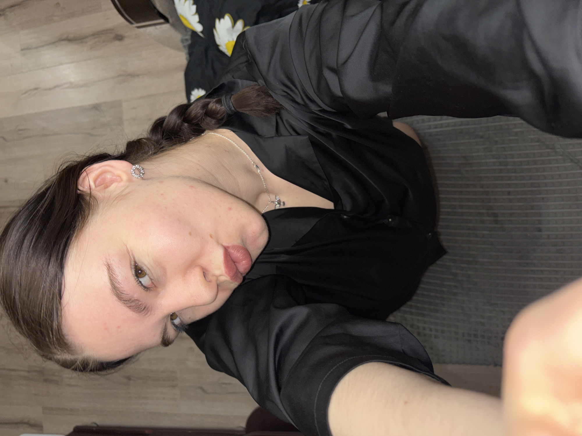 A young woman with light skin and brown hair braided to the side is depicted in a selfie-style photograph. She has light-colored eyes and is wearing a silver necklace with a charm. She is wearing a dark-colored, possibly satin, garment. She has an earring in her left ear. The background includes what appears to be a light wood floor, a gray striped surface, and a floral patterned fabric. The picture does not explicitly identify the city of Perm, Russia, but provides no evidence to the contrary.