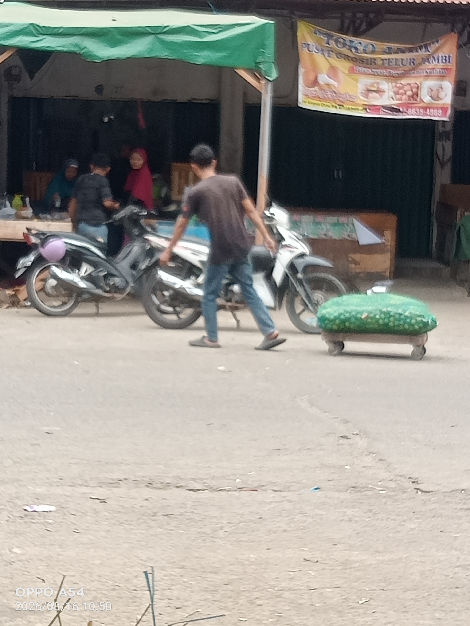A medium shot captures an outdoor scene in Jambi, Indonesia, focusing on a man in a dark t-shirt and blue jeans pushing a cart laden with green produce. The cart, a low-lying platform on wheels, is covered with a green net, and the produce appears to be small and round, possibly limes or small melons. The man is in motion, his left leg extended forward as he propels the cart. Behind him, two motorcycles are parked. The first motorcycle, a dark-colored scooter, is on the left, and a white motorcycle is partially visible to its right. A purple object, possibly a helmet, is attached to the back of the dark scooter. In the background, under a green awning, there are several people visible inside a building. One person in a red head covering is prominent, and two others are seated. A yellow banner with Indonesian text and images of eggs is hung above the storefront. The ground is paved with asphalt. The overall impression is a busy street scene in a commercial area, likely a market or roadside stall.