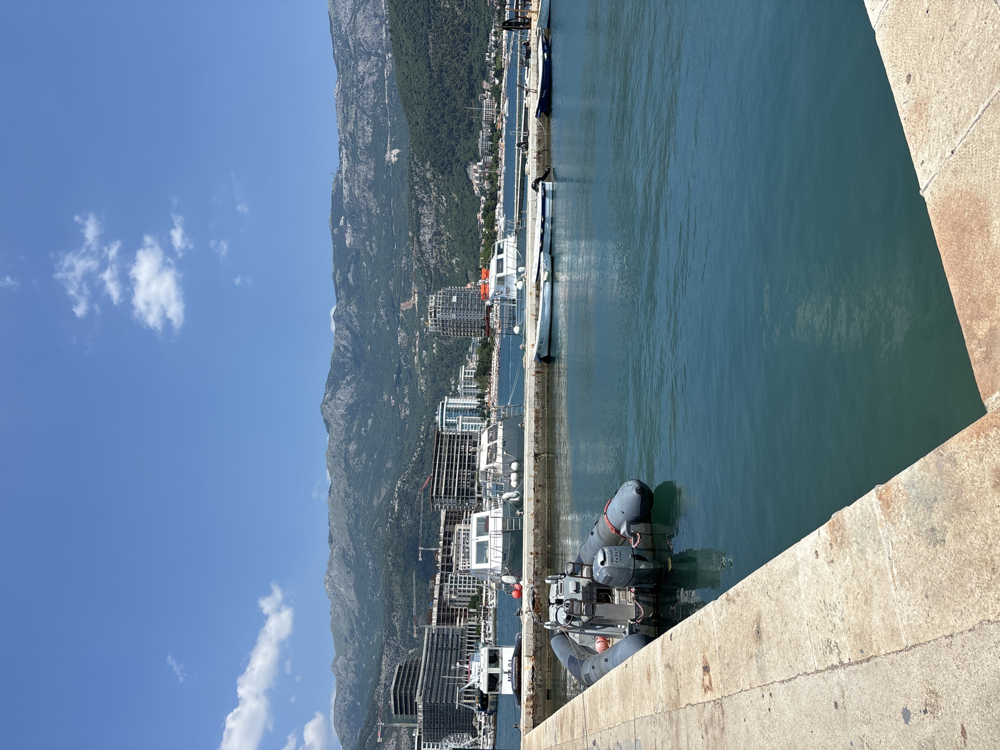 The image depicts a sunny day at a harbor in Budva, Montenegro. The scene is dominated by a calm, turquoise body of water reflecting the bright blue sky and a few scattered clouds. In the foreground, a concrete pier juts out into the water, with a rigid inflatable boat (RIB) with an outboard motor docked at its edge. The RIB is gray with black inflatable tubes and has red accents.
Beyond the water, a cityscape of modern buildings, some under construction, lines the shore. Behind the city, a lush, green mountain rises steeply, its slopes dotted with trees and some visible rocky outcrops. Several other boats, including a white passenger ferry with orange life vests visible, are moored along the harbor. The sky is clear and bright, suggesting midday. The overall impression is one of a bustling coastal town in warm weather. No people are clearly visible, but the presence of the boats and the developed shoreline indicates human activity.