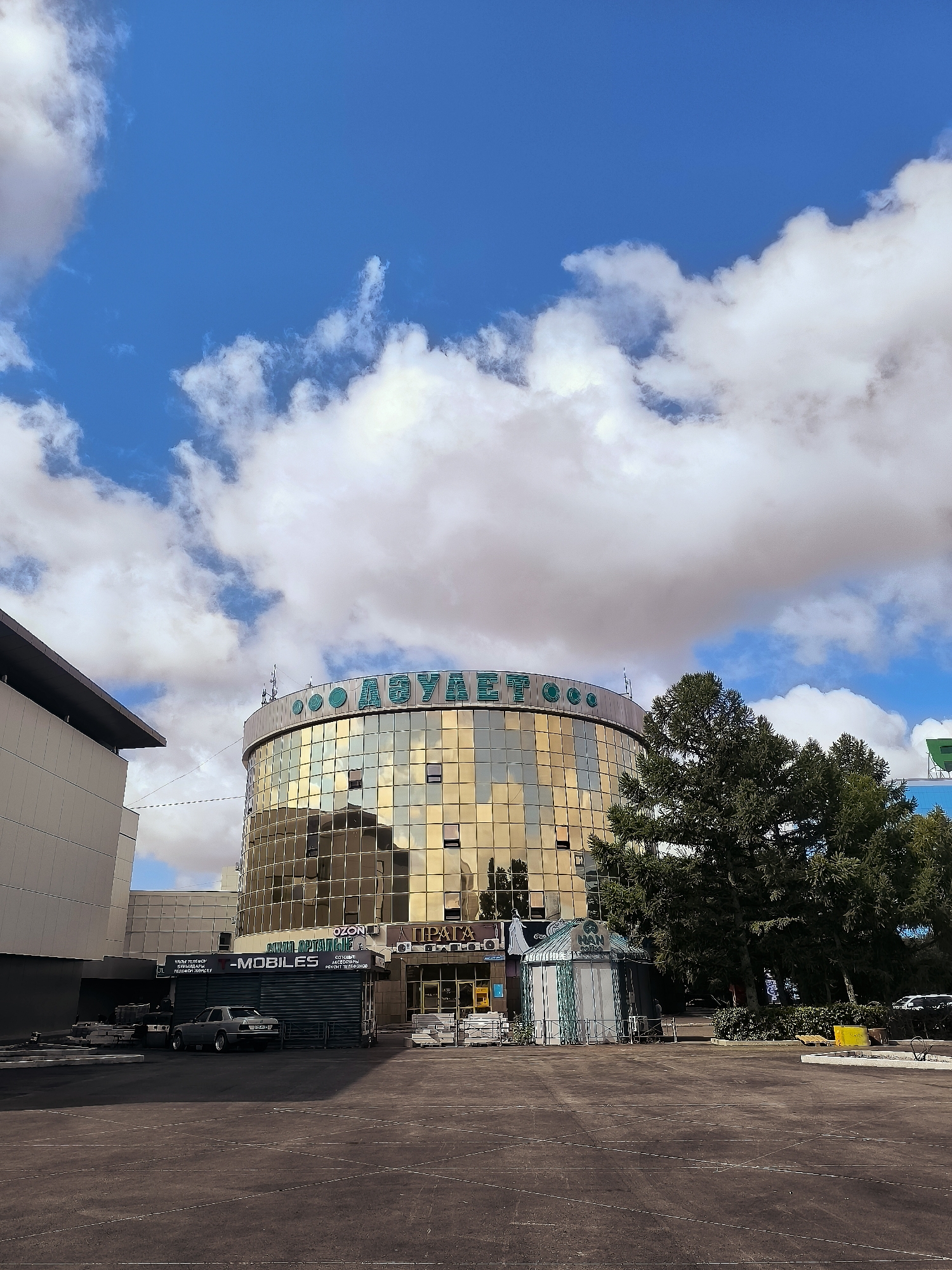 The image captures a low-angle view of a modern, cylindrical building with a golden, reflective facade, located in Qarağandı city administration, Kazakhstan. The building is topped with a green sign that reads "ДОЦУАЕТ" in Cyrillic letters. In front of the building, there is a dark, paved area, possibly a parking lot, with white lines painted on it. To the left, a portion of another building with a flat, grey facade is visible. To the right of the main building, a large, mature tree with dark green foliage stands prominently. A small, striped kiosk with green and white stripes is situated in front of the tree, near the main building. A dark-colored sedan is parked in front of a green, shuttered storefront labeled "-MOBILES". The sky above is a bright blue with scattered white clouds. The overall scene depicts an urban environment on a clear day.