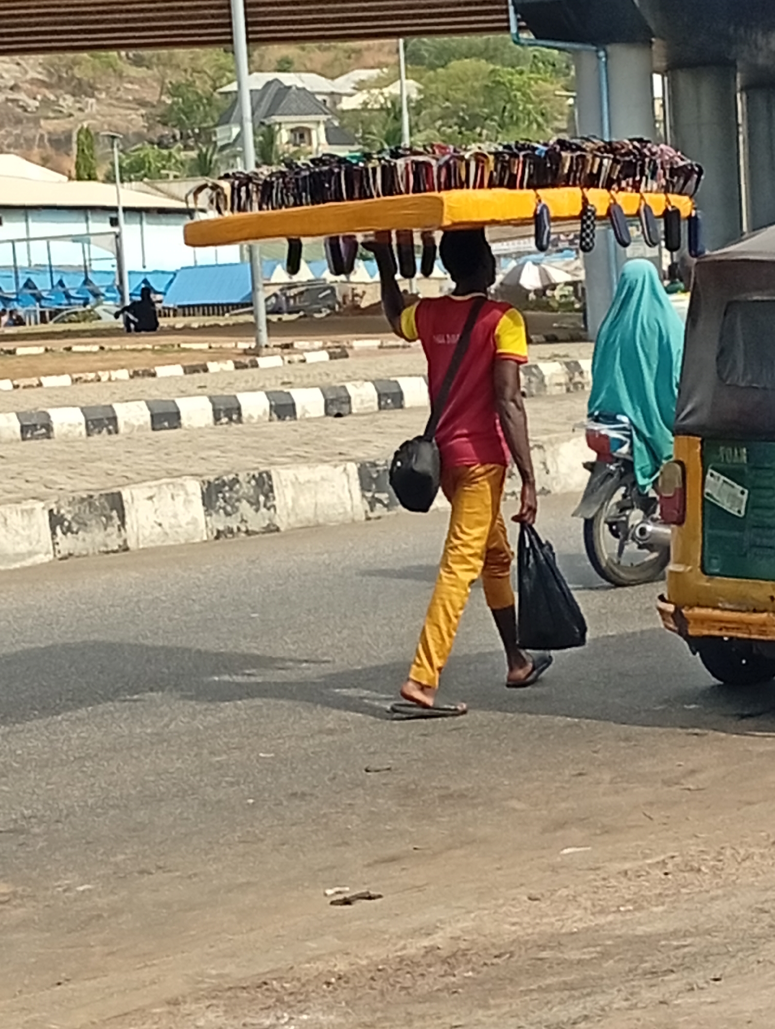 In Lokoja, Nigeria, an adult male carries a yellow tray laden with numerous packaged items on his head while walking along a paved road. He wears a red and yellow shirt, yellow pants, and flip-flops. A black bag hangs from his shoulder, and he carries another black bag in his hand. To his right, a woman in a blue hijab sits on a motorcycle, and a green and yellow auto-rickshaw (tricycle) is partially visible. In the background, there are buildings and a person sitting on the ground, along with a black and white striped curb. The scene appears to be outdoors under a bridge, in daylight.